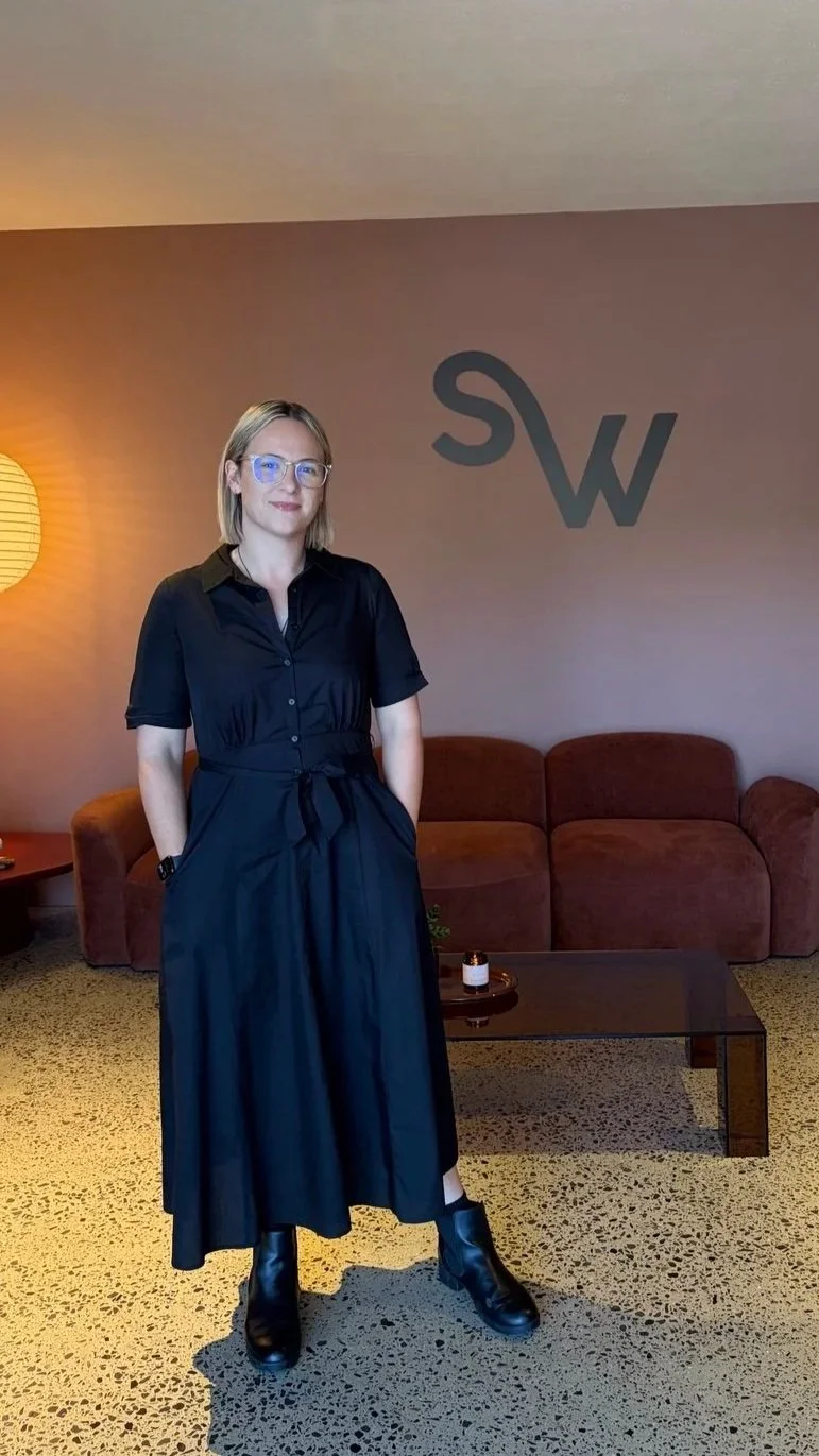 A woman with blonde hair and glasses stands in a living room, wearing a black dress and boots. Behind her is a brown sofa, a coffee table with a candle and small plant, a lamp on a side table, and a wall with a large black logo consisting of the letters "S" and "W."