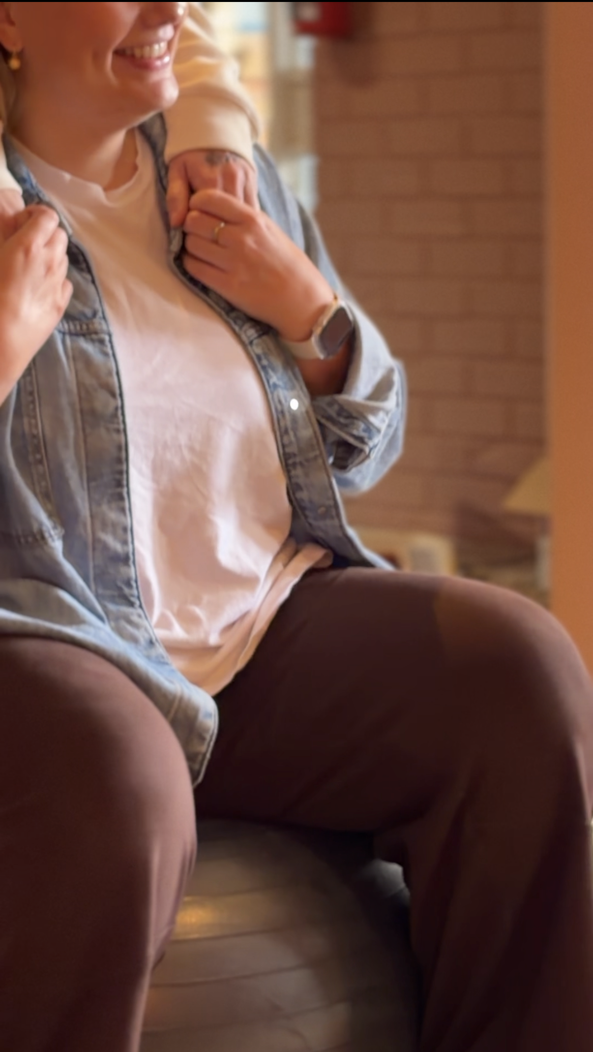 Close-up of a woman smiling, wearing a light pink T-shirt and a denim jacket, sitting on a brown leather chair, with an arm over her shoulder.