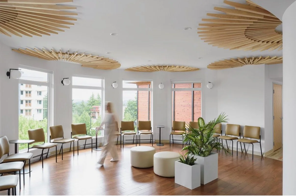 A waiting room with white walls, large windows, and wooden ceiling accents shaped like sunbursts. There are multiple beige chairs arranged along the perimeter, a woman walking through, and potted plants in white containers.