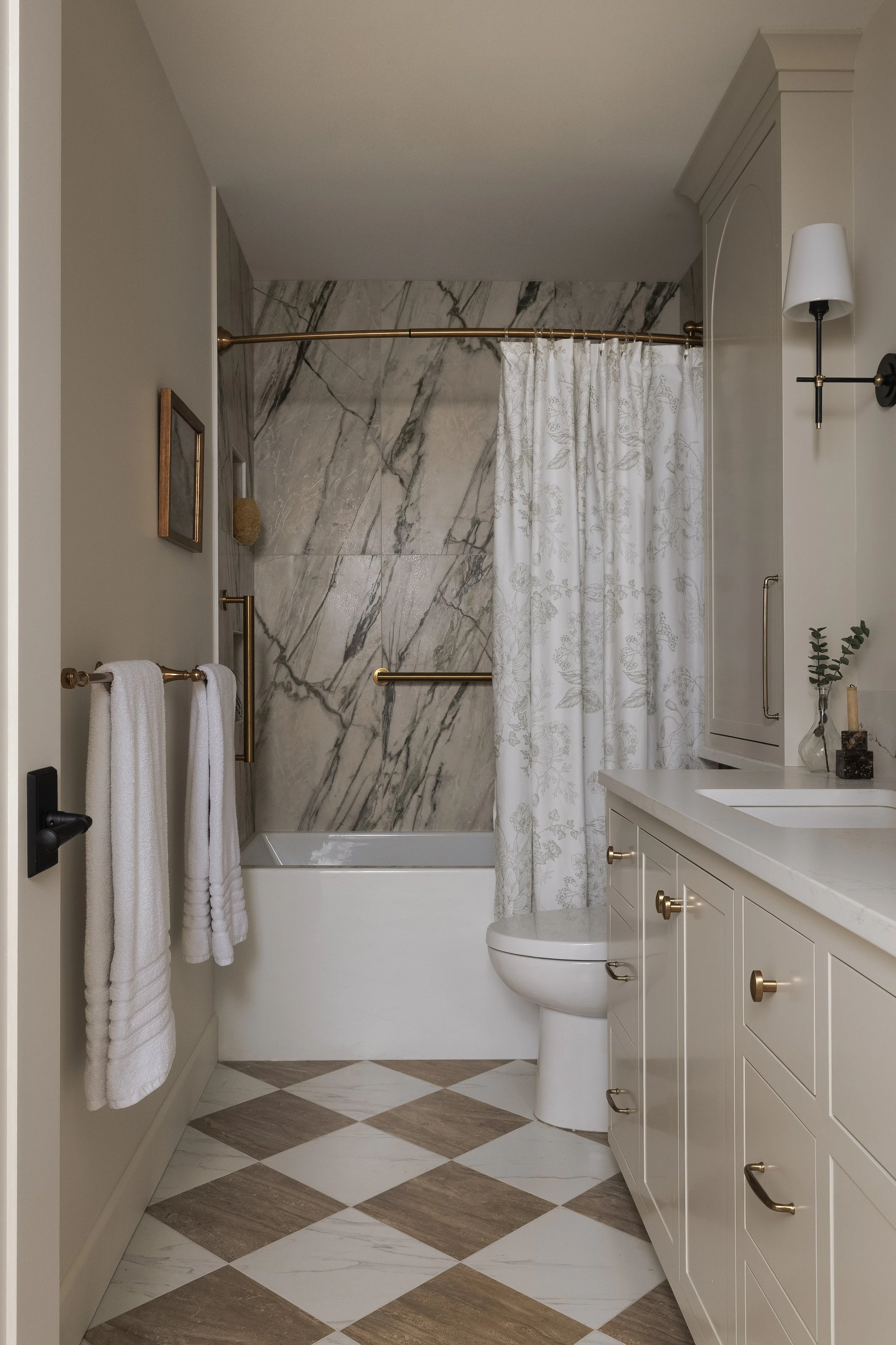 A modern bathroom with a white vanity, marble tile walls in the shower area, and a matching shower curtain. Two white towels hang on a gold towel rack, and there is a toilet next to the vanity. The floor has a brown and white checkered pattern.