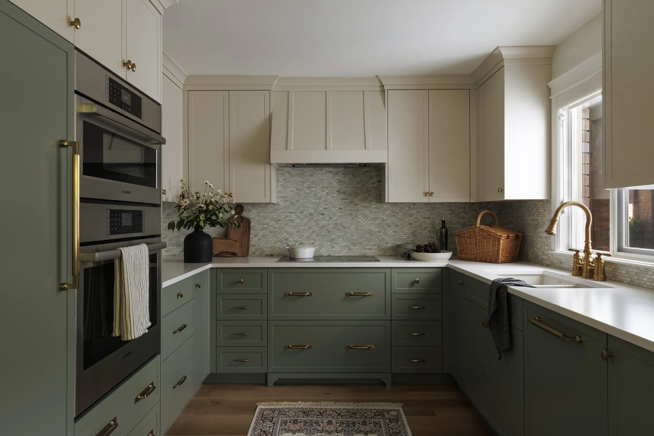 Kitchen with green lower cabinets and white upper cabinets, stainless steel oven, window with light streaming in, and decorative items on the counter.
