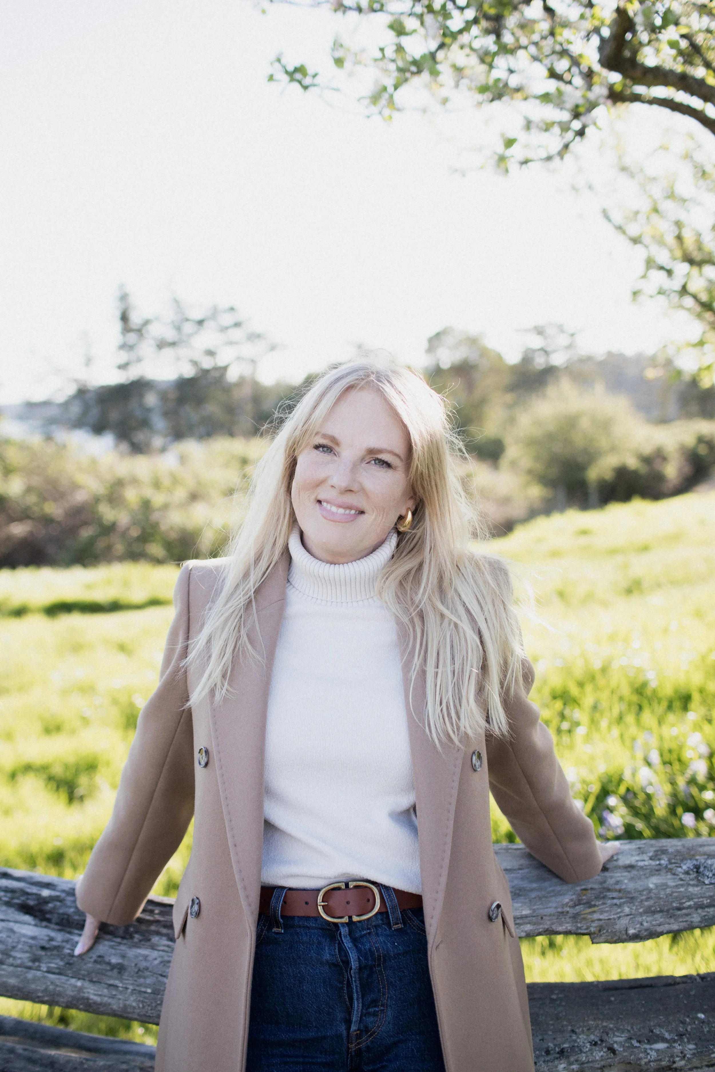 Woman with blonde hair smiling outdoors in a park or garden with green grass and trees, wearing a beige coat over a white turtleneck, standing next to a wooden fence.