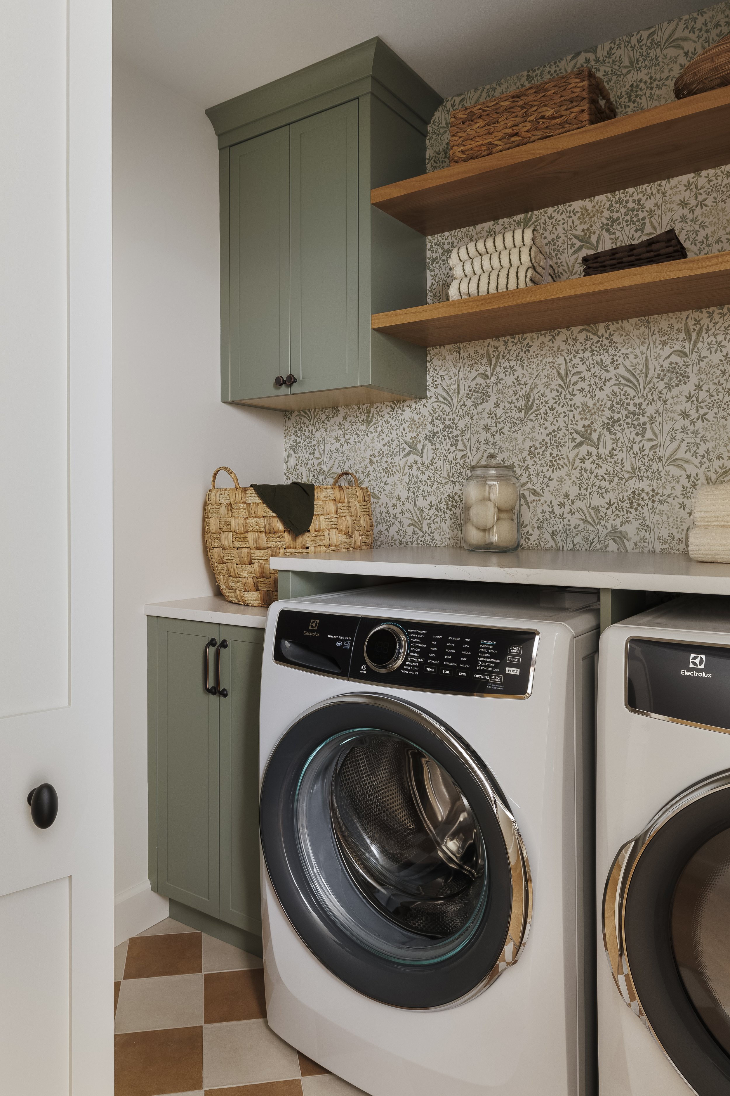 Cottage Laundry room with green cabinets in Sherwin Williams Evergreen Fog, terracotta checkerboard floor, floating wooden shelves for storage with folding counter top. Kitchen Interior Design in Victoria BC