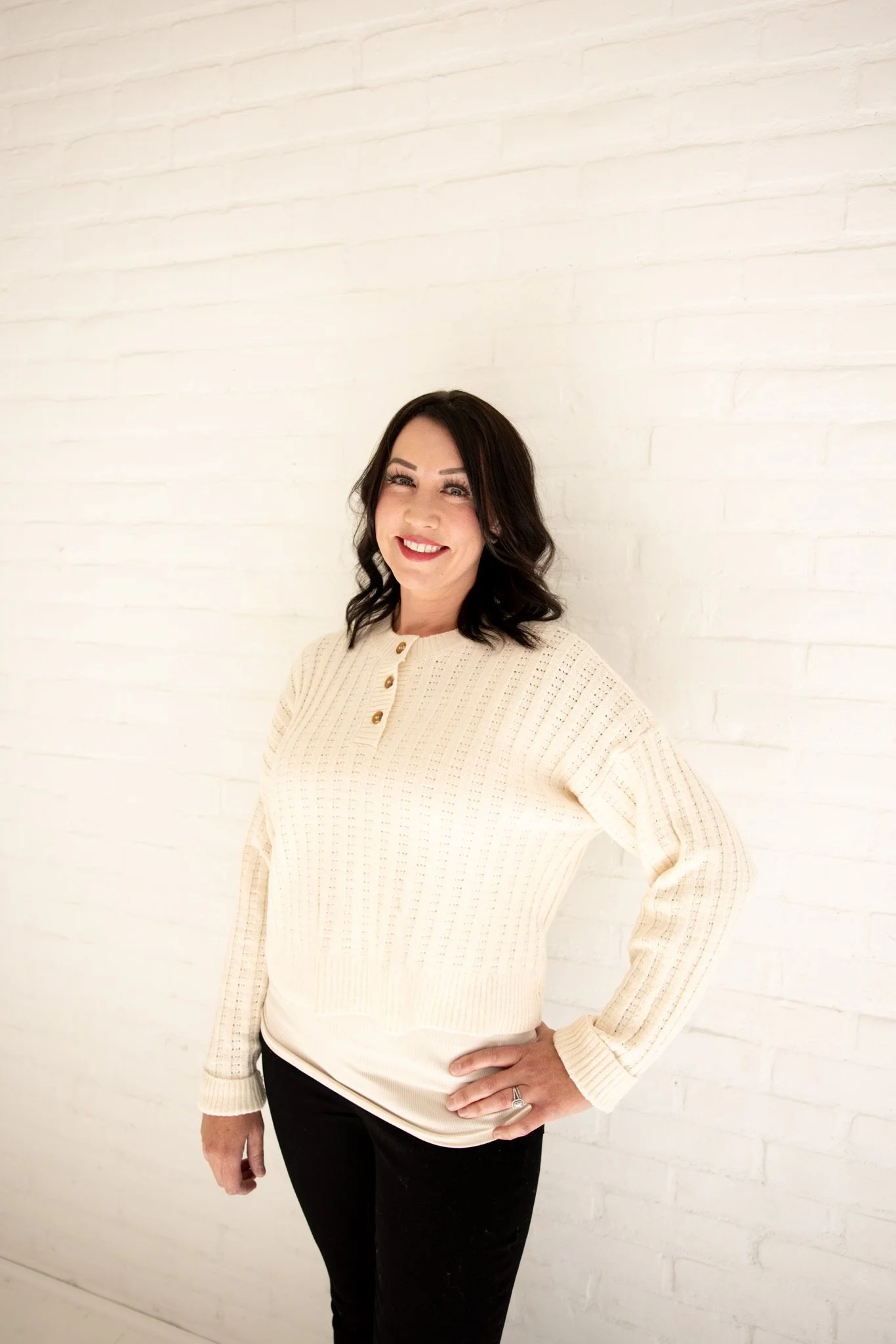 Woman with dark wavy hair wearing a cream knit sweater with gold buttons and black pants, smiling and standing against a white brick wall.