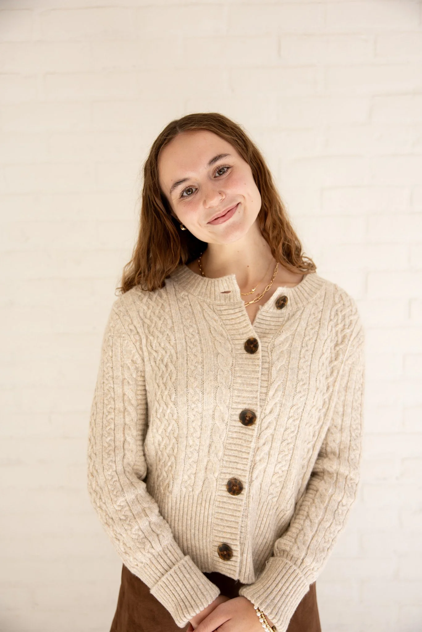 A young woman with wavy brown hair wearing a beige cable knit sweater with large brown buttons, gold jewelry, and a brown skirt, standing against a white brick wall, smiling softly.