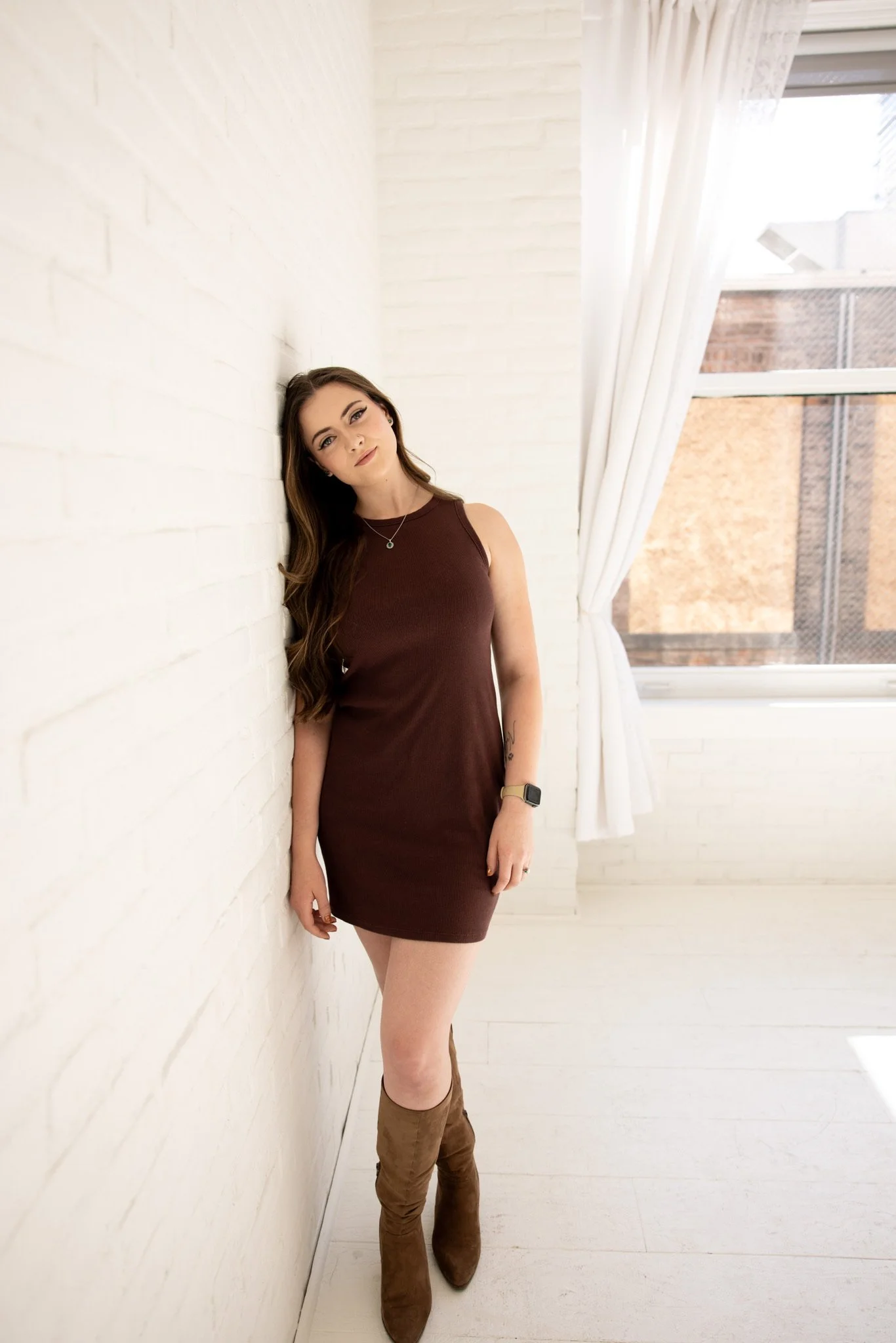 Young woman with long brown hair wearing a sleeveless maroon dress and brown boots, standing in a bright room with white brick walls and a large window with sheer curtains.