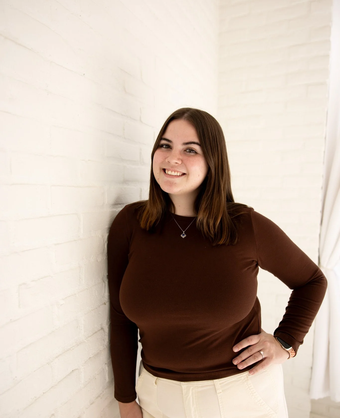 A woman with brown hair, wearing a brown long-sleeve shirt and beige pants, smiling and standing against a white brick wall indoors.