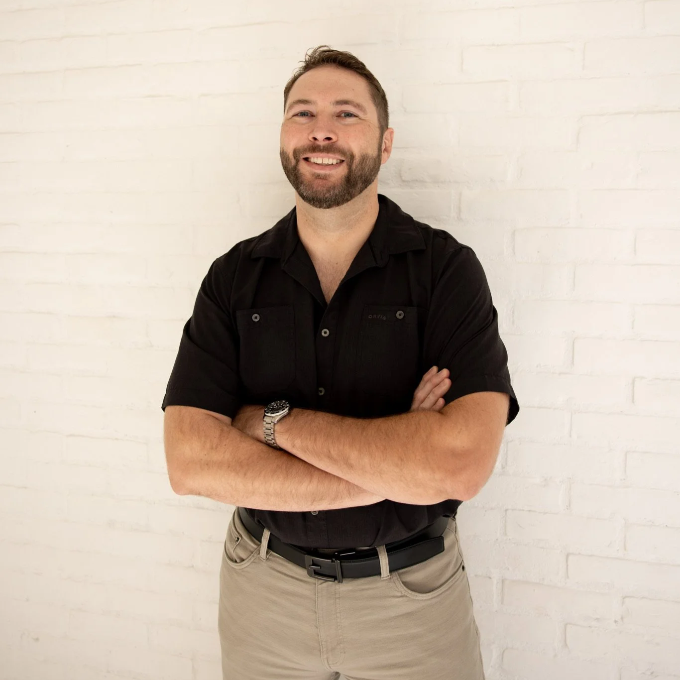 A man with a beard and short hair smiling, standing with arms crossed in front of a white brick wall, wearing a black short-sleeve button-up shirt, beige pants, and a watch.
