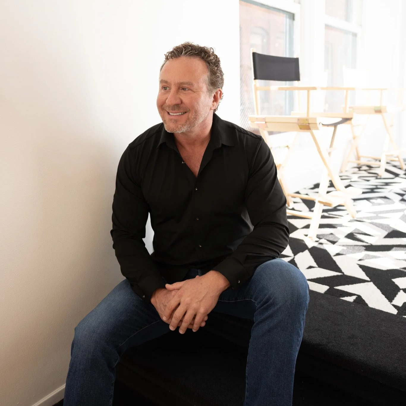 Smiling middle-aged man with curly hair and a beard, wearing a black shirt and blue jeans, sitting on a black cushion in a brightly lit room with a patterned black and white rug in the background.