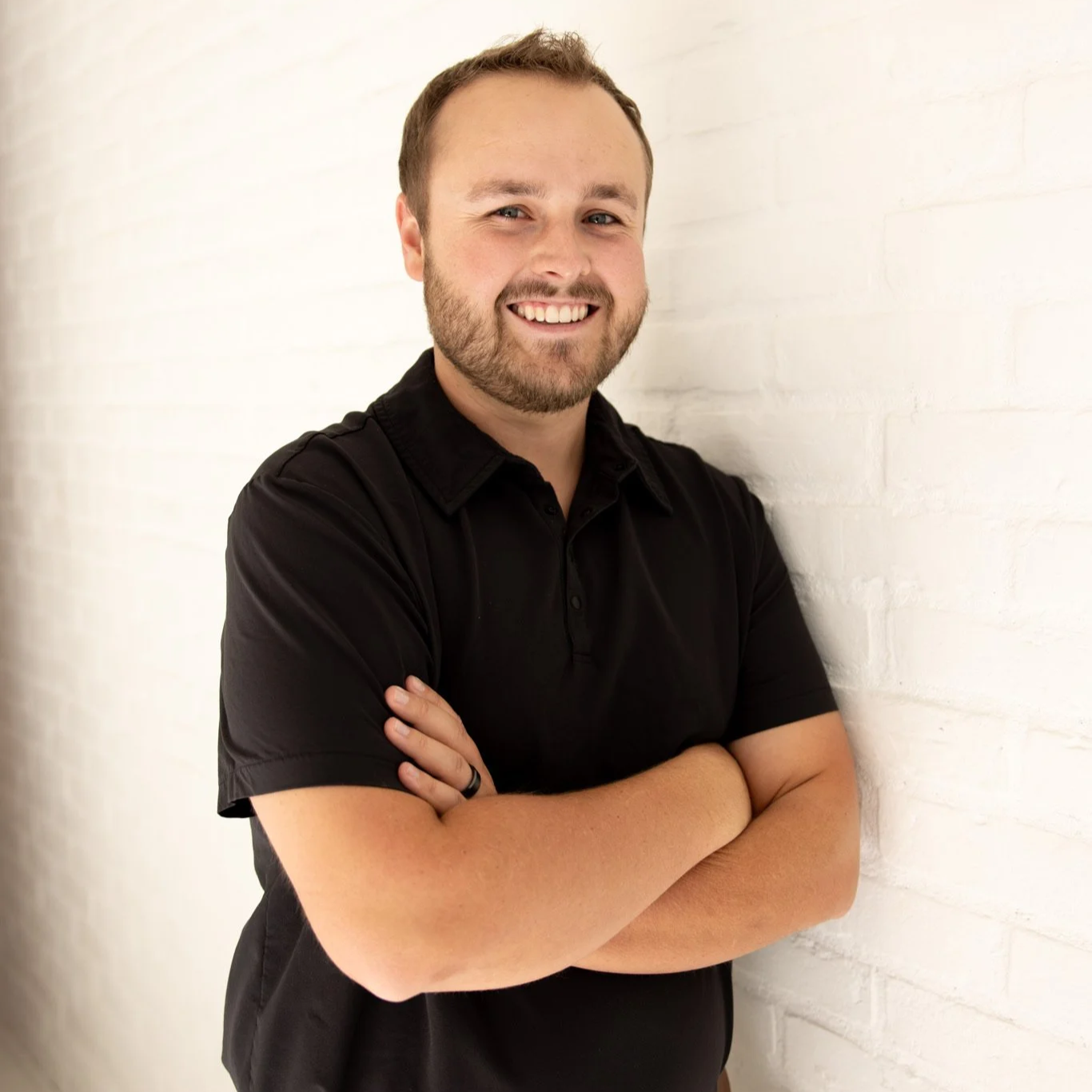 A man with a beard and short hair smiling, wearing a black polo shirt, crossing his arms, standing against a white brick wall.