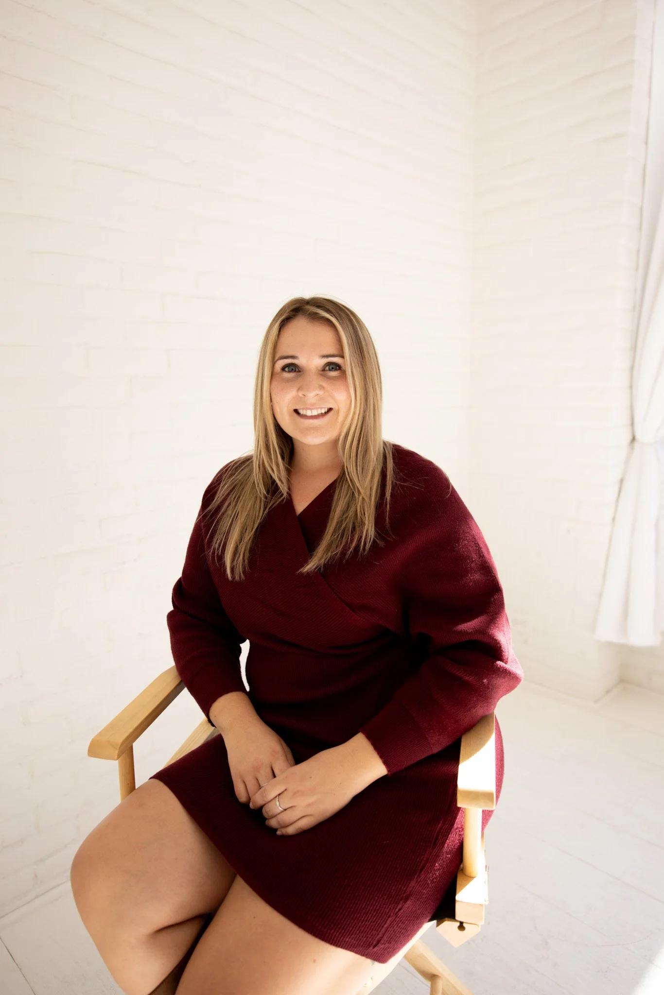 A woman with long blond hair wearing a burgundy dress sitting on a wooden chair in a well-lit room with white brick walls and a window with white curtains.