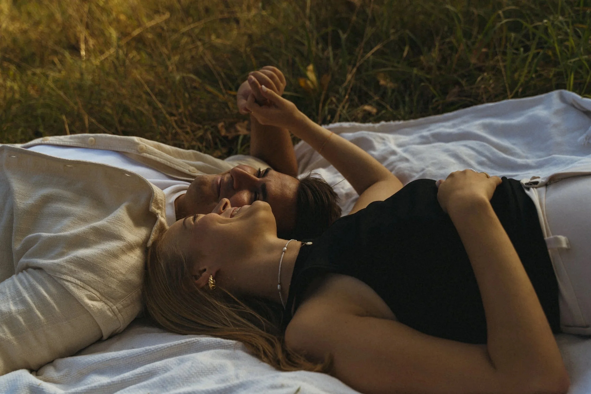 Zwei Frauen liegen eng umarmt auf einer Decke im Freien, scheint spät am Tag oder bei Sonnenuntergang, umgeben von Gras.