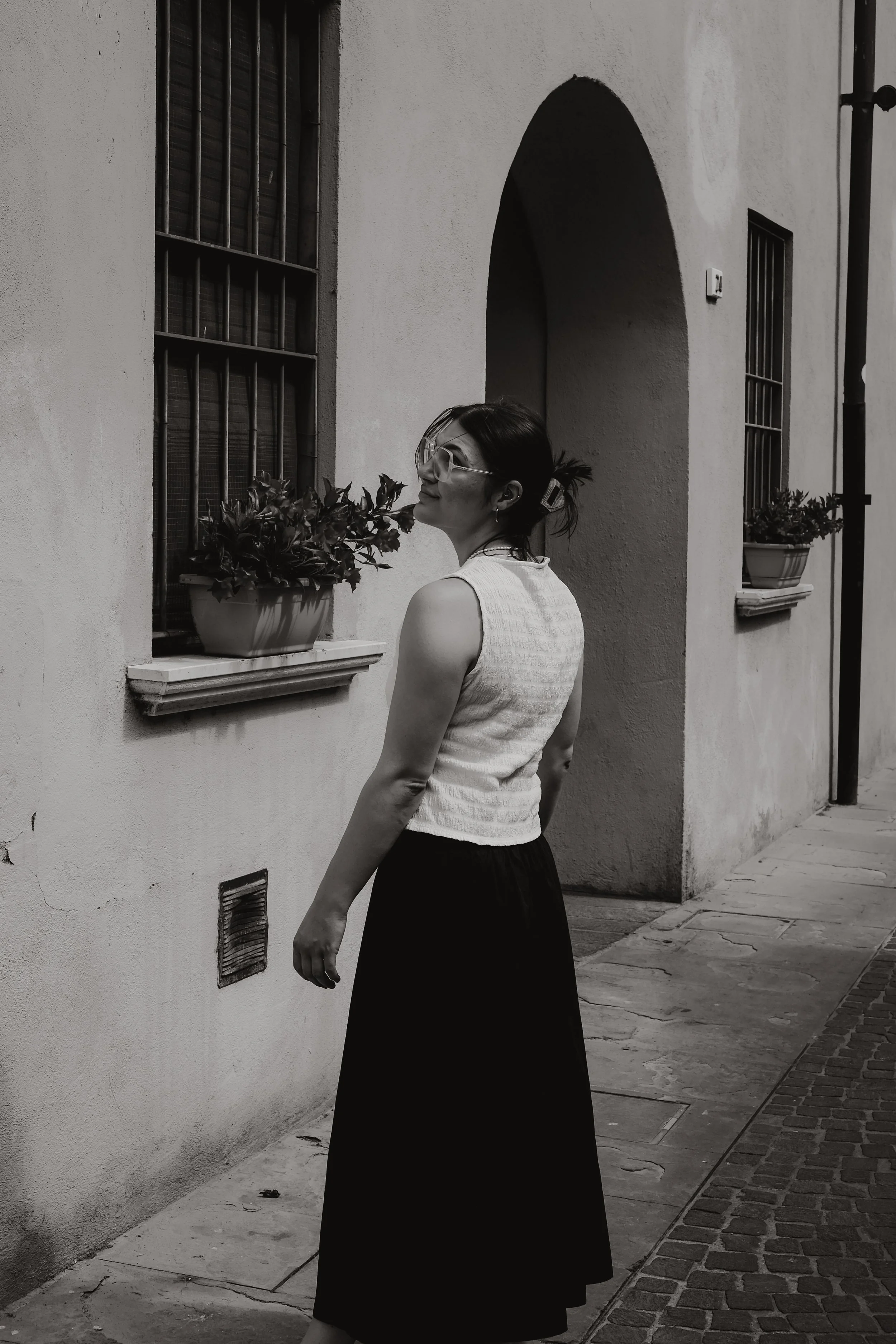 Schwarz-weiß-Foto einer Frau mit dunkelgelbem Hemd und dunklem Rock steht an einer Hauswand mit Fenstern und Pflanzenschalen, die mit Blumen gefüllt sind.