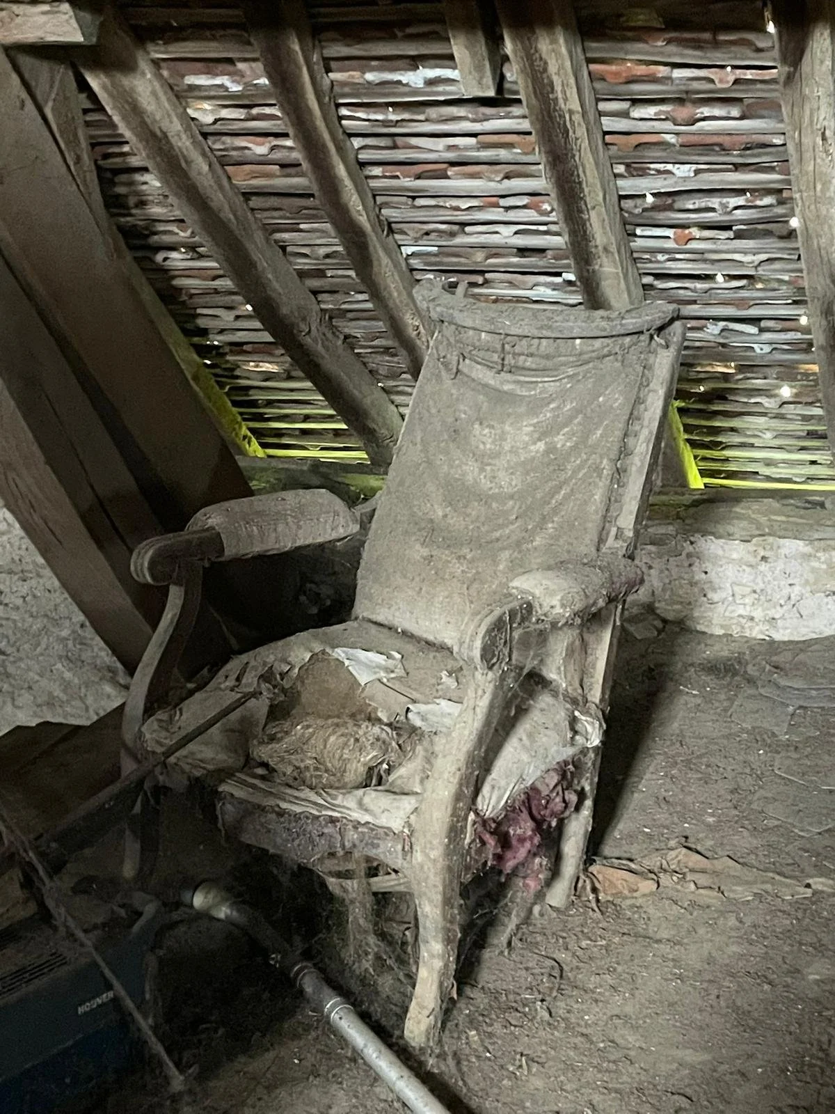 A dusty, old wooden chair with worn armrests and a tattered seat sits in a shed. The shed has a thatched roof with overlapping wooden slats and rough, uneven dirt floor.