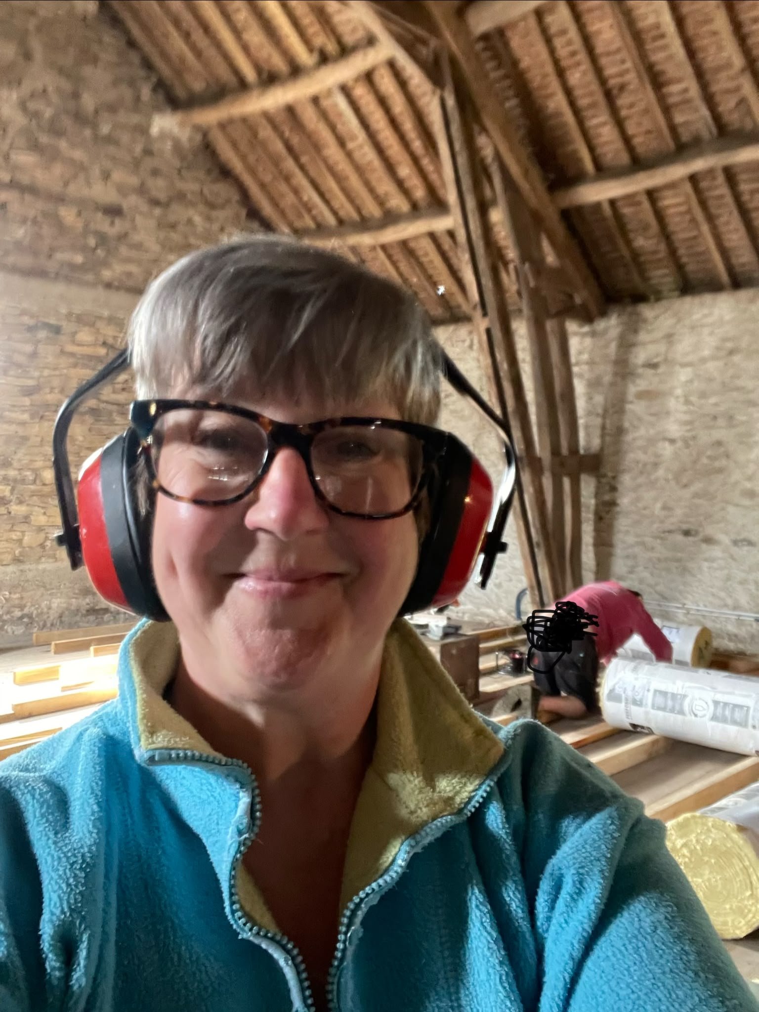 A person wearing glasses and a hearing protection mask, smiling at the camera inside a rustic barn with wooden beams and brick walls.