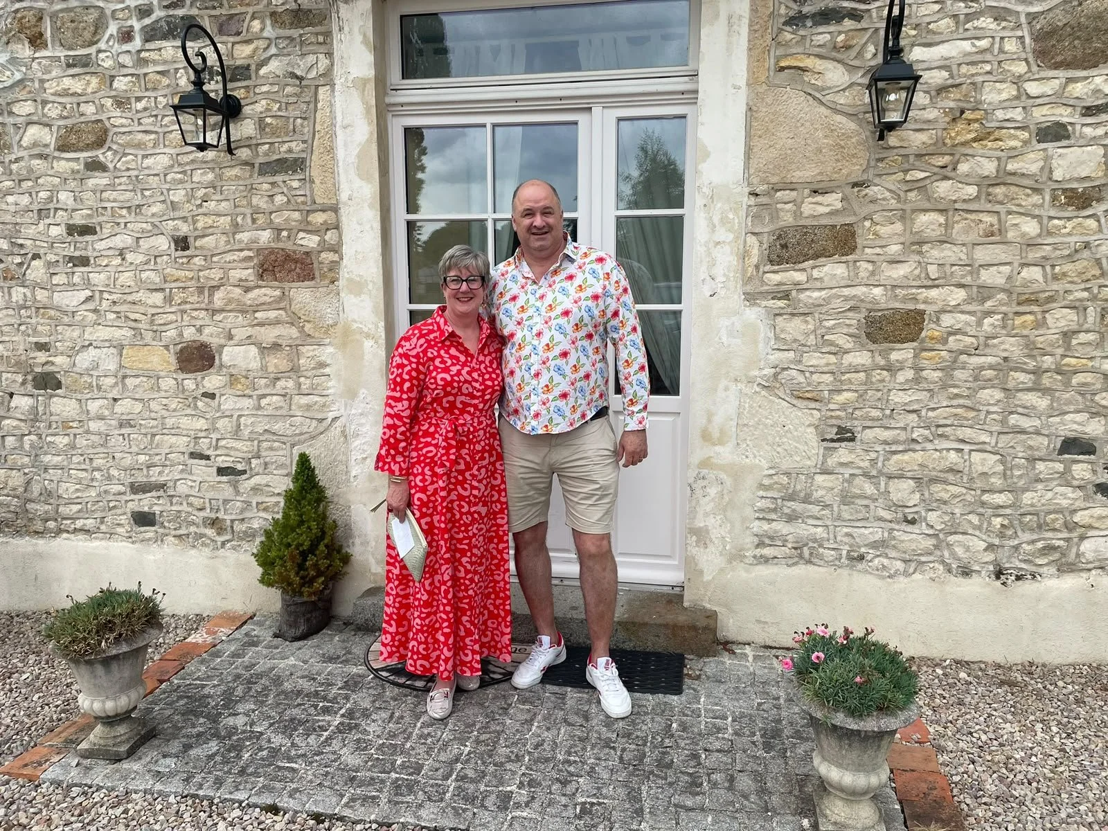 Two people standing in front of a stone house door, smiling. The woman is wearing a red dress with a white pattern, and the man is wearing a colorful, patterned shirt and beige shorts. There are potted plants on either side of them.