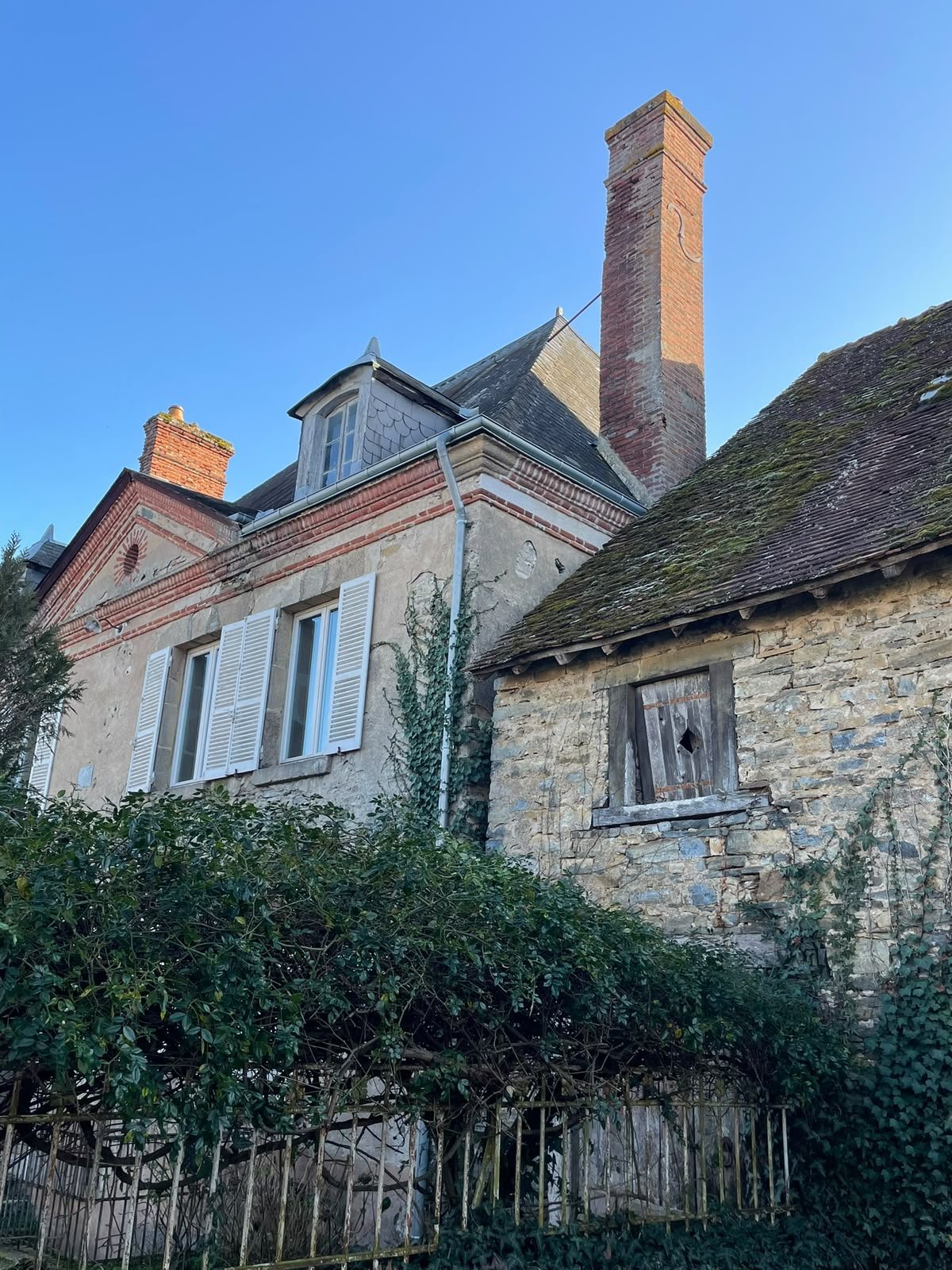 The image shows an old stone house with a sloped roof and a small, boarded-up window. Next to it is a larger house with beige walls, open white window shutters, a chimney, and a small dormer window on the roof. Ivy is growing on the side of the larger house, and there are bushes in the foreground. The sky is clear and blue.