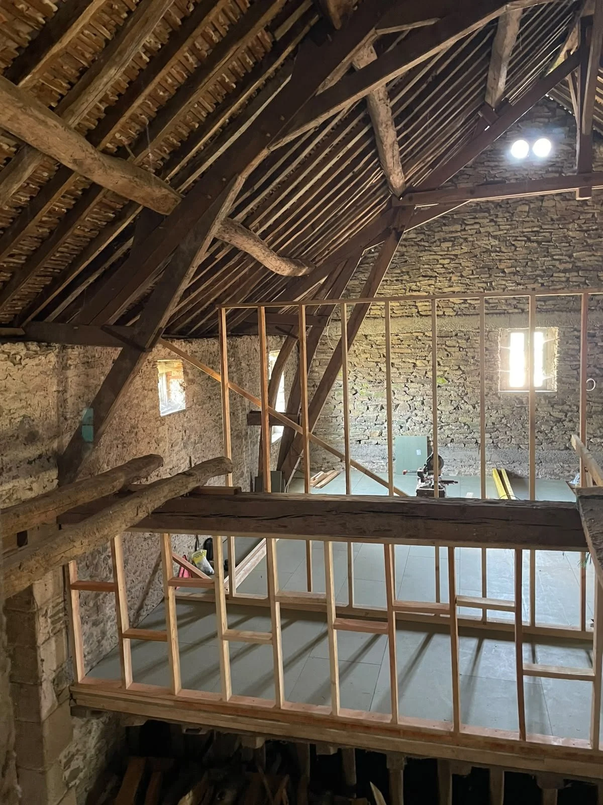 The image shows the interior of an old stone building under renovation, with recently constructed wooden framing for a new wall or floor. The roof is made of wooden beams and tiles, with some rustic, weathered beams visible. There are small windows on the stone wall allowing sunlight to enter, and construction tools can be seen on the floor.