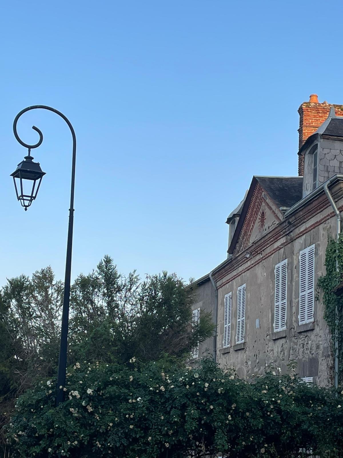 A vintage street lamp on a curved pole standing beside an old stone building with white shutters and ivy, with trees and a clear blue sky in the background.
