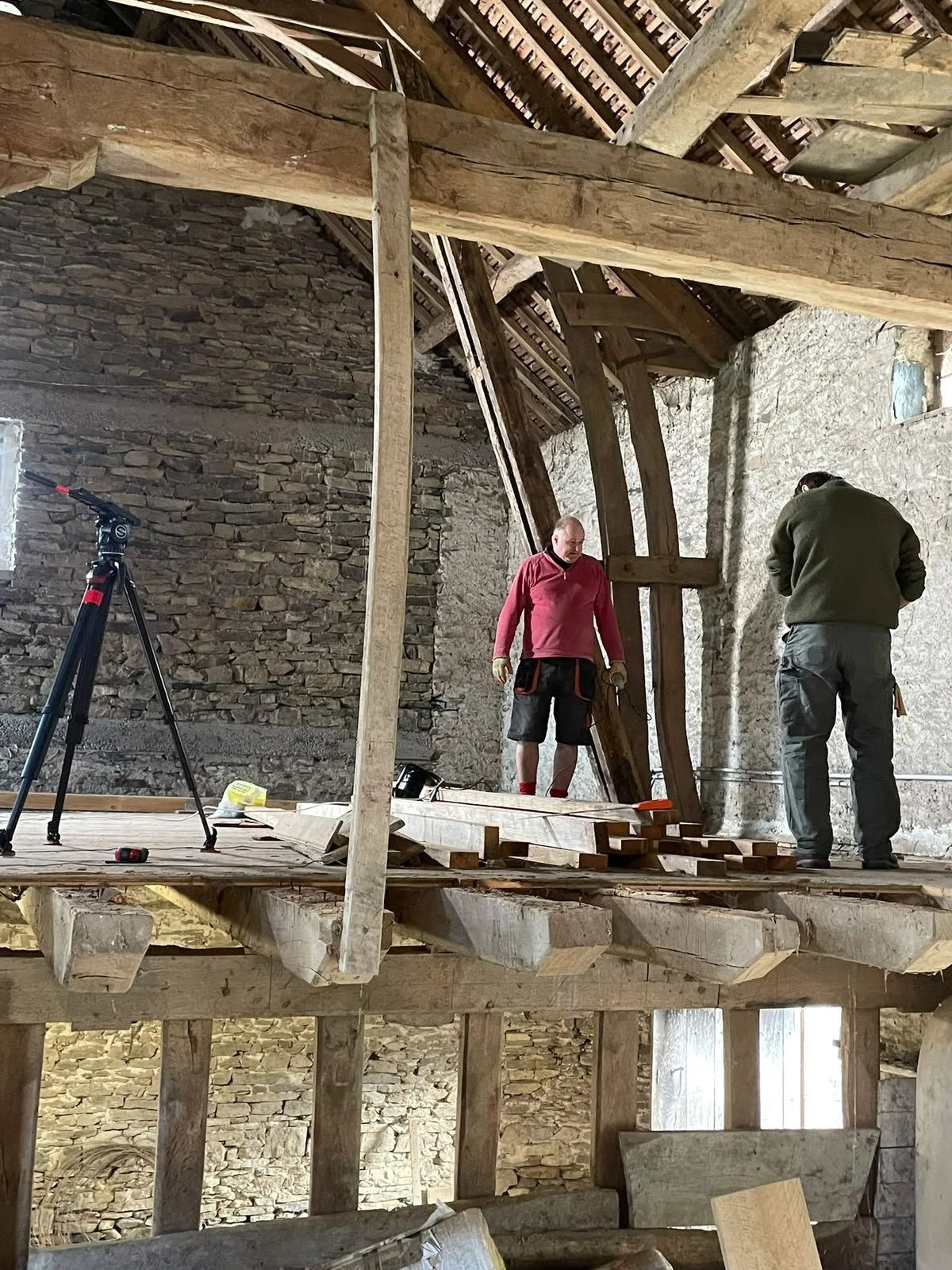 Two men are working on a wooden construction inside a rustic stone building. One man in a red shirt appears to be overseeing or directing, while the other in a dark sweater is focused on a task. Construction tools and materials are scattered around, 