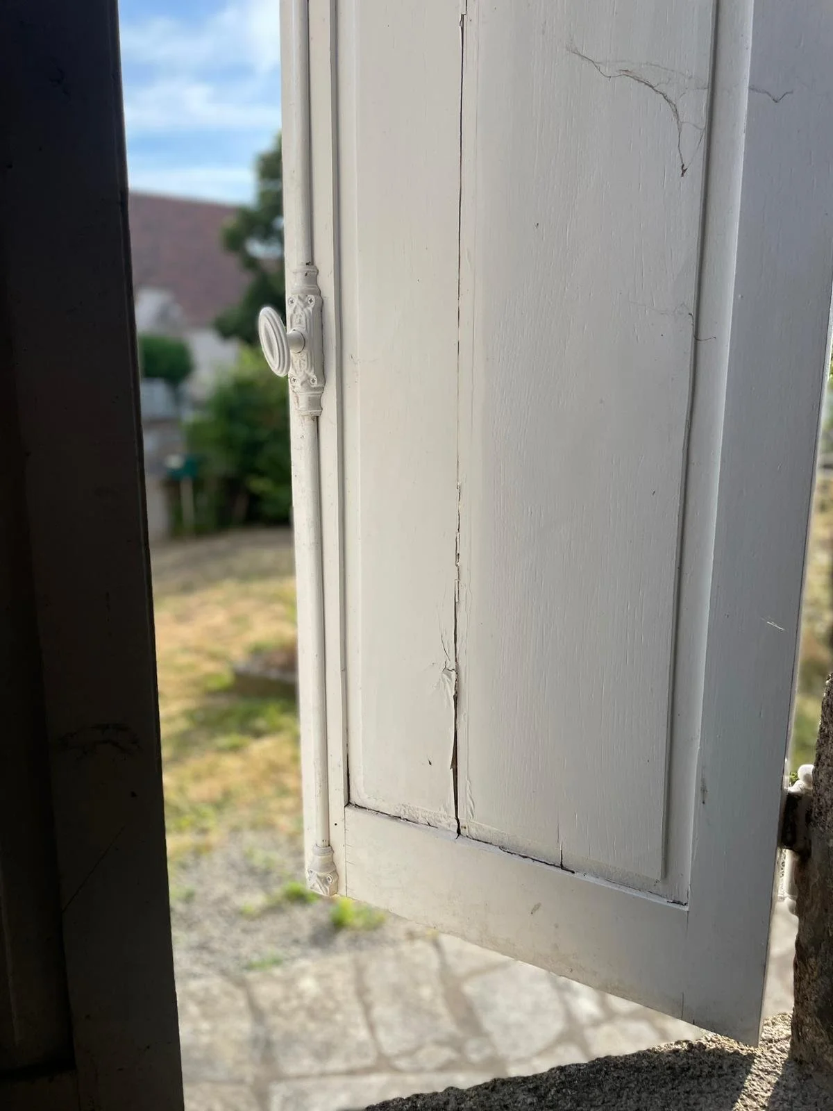 Close-up of a small white wooden door with a crack running vertically through the middle, slightly open, revealing a blurry outdoor scene with trees and houses.