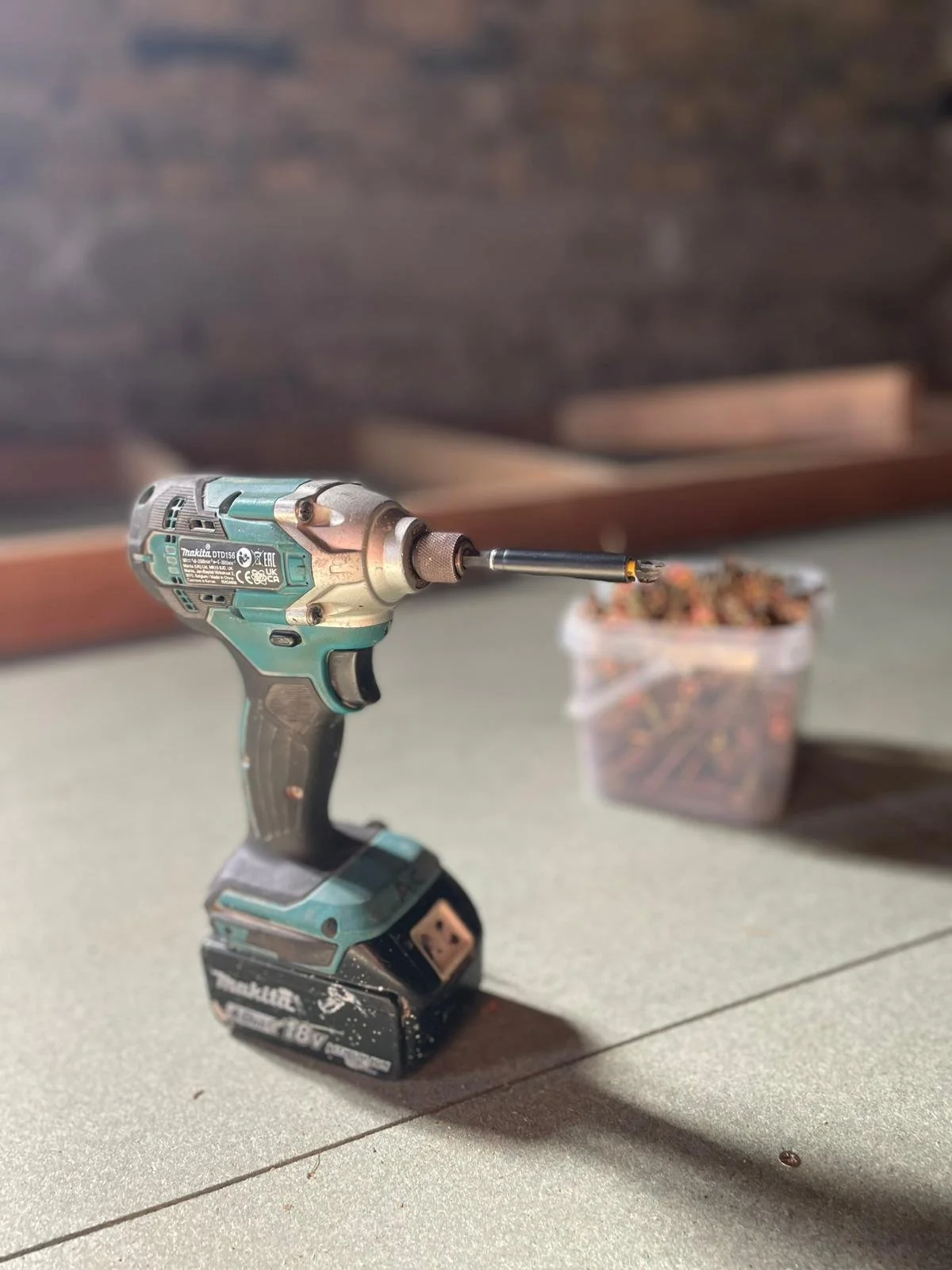Electric drill on a gray surface with a container of screws in the background.