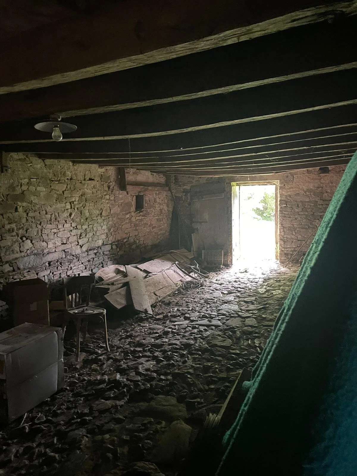 Interior of an old stone building with a dirt and stone floor, cluttered with debris and broken furniture, illuminated by sunlight coming through an open door.
