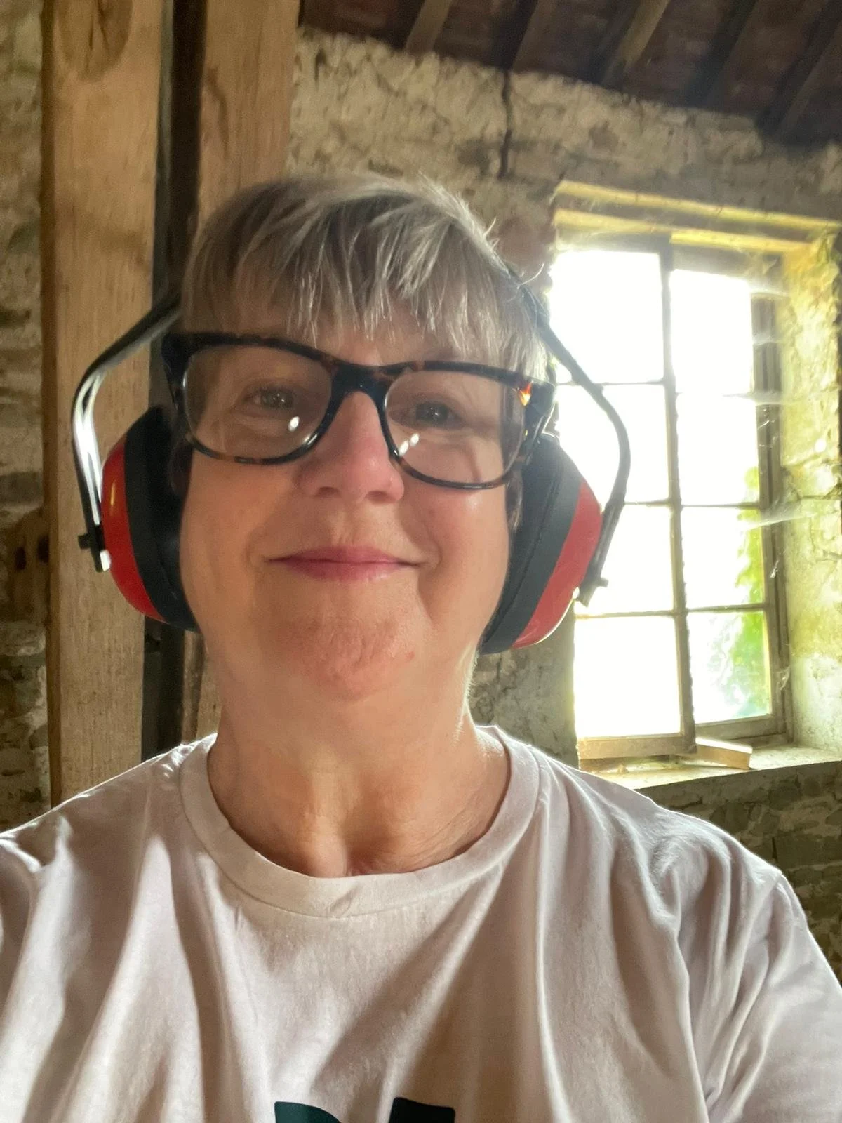A person with short gray hair wearing large safety glasses and red earmuffs, smiling in a rustic room with stone walls and a window in the background.