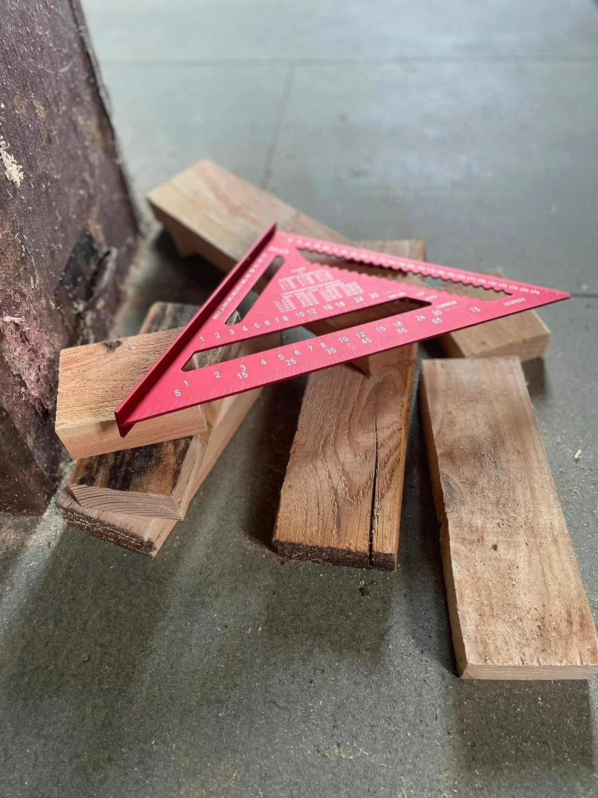 A red T-square ruler resting on several pieces of wooden planks in a workshop or construction area.