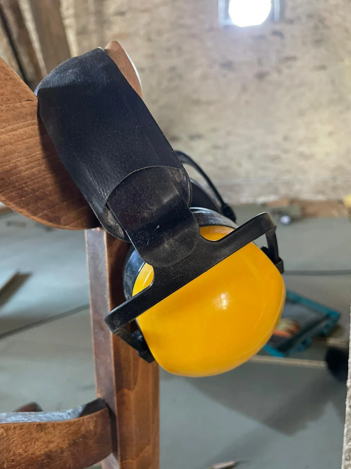 A yellow mug hanging from a black mug holder attached to a wooden part of furniture or rack, with a black strap around the top of the mug. The background includes a rough textured wall and a ceiling with a bright light source.