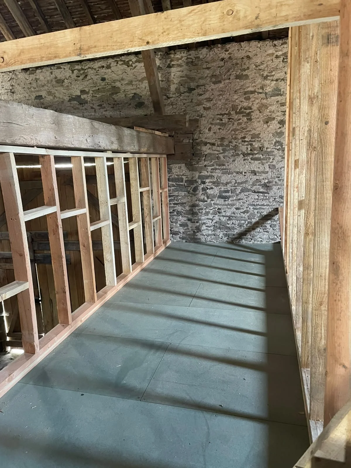 Interior view of an unfinished room with exposed stone wall, wooden framing, and concrete floor, with sunlight casting shadows on the floor.