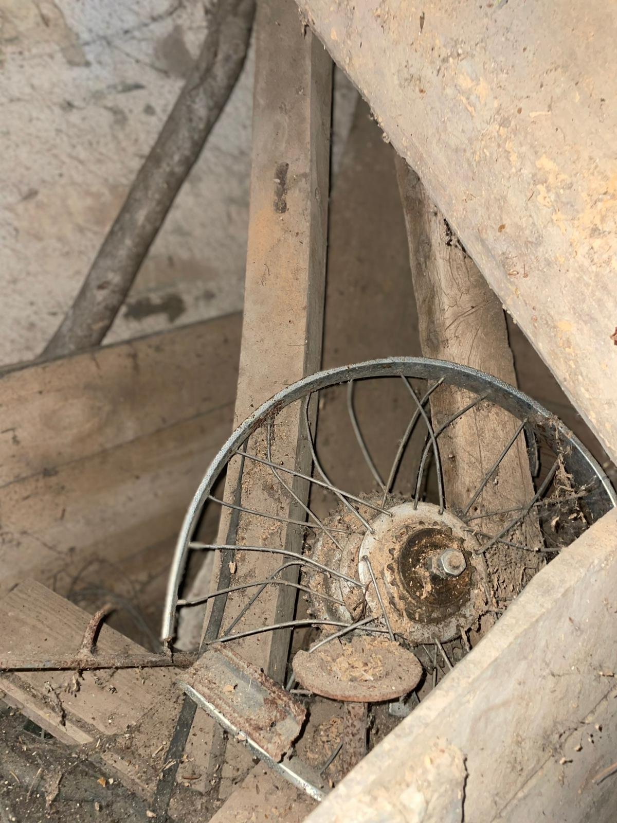 A dusty, old metal wheelbarrow with chipped paint, lying on a dusty wooden floor with a piece of rusted metal and wood debris around.