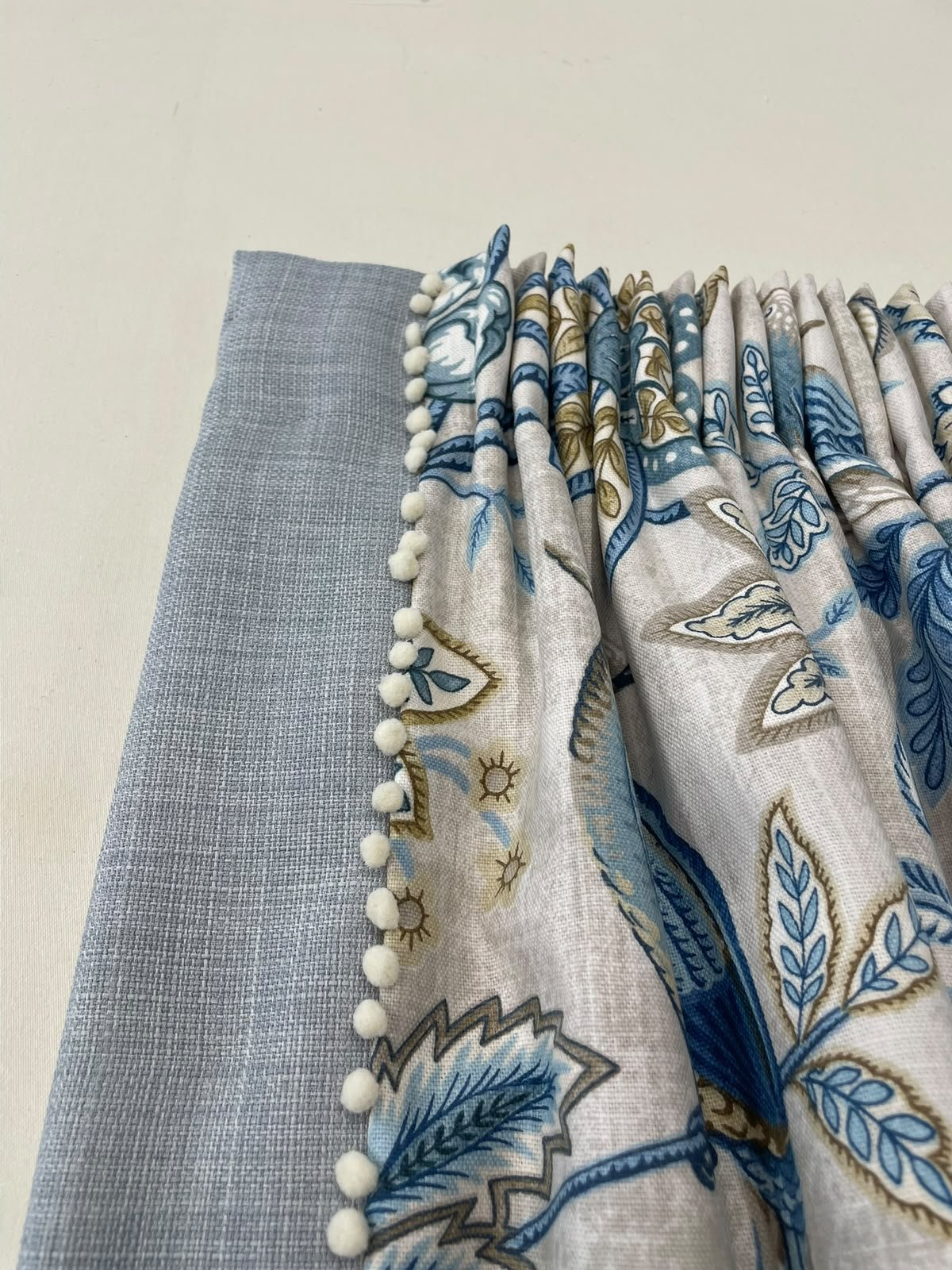 Close-up of a curtain with a light blue, patterned outer layer decorated with white pom-pom trim and a solid, textured light gray inner curtain, on a white background.