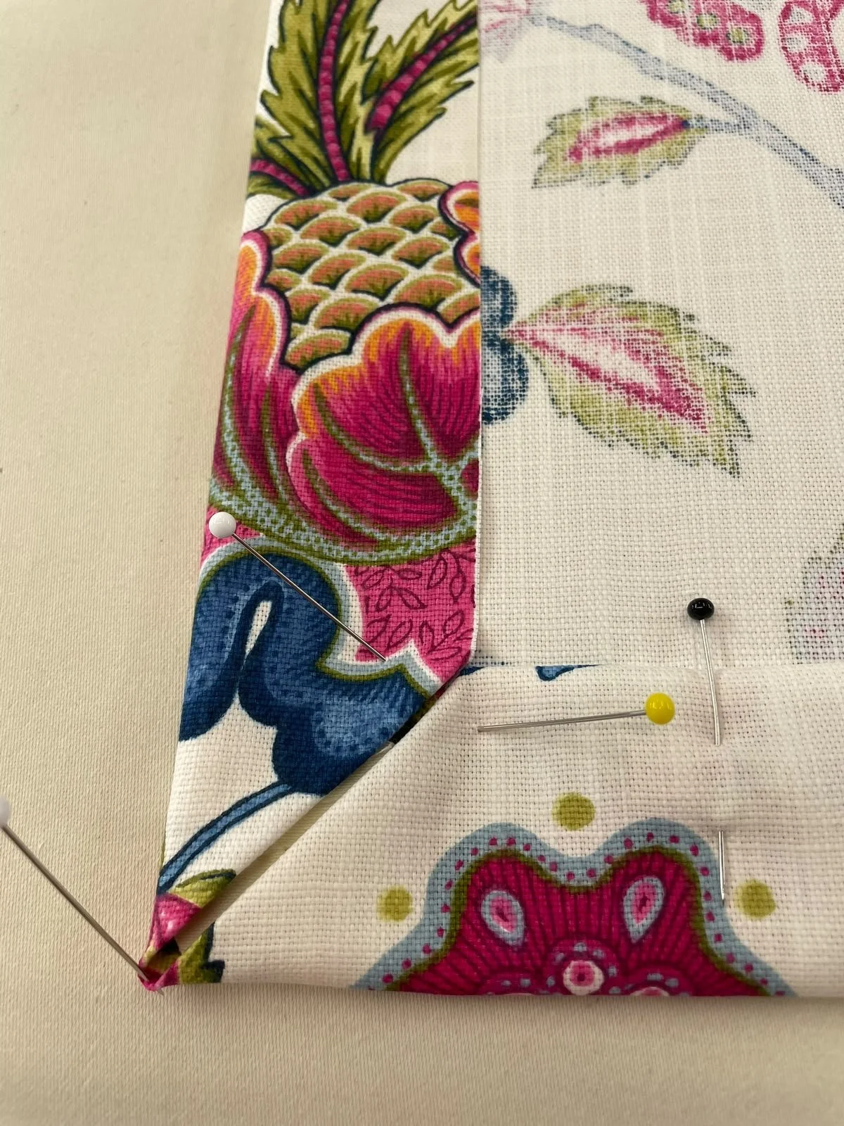 Close-up of fabric with bright floral and fruit patterns, pinned with sewing pins on a light-colored surface.