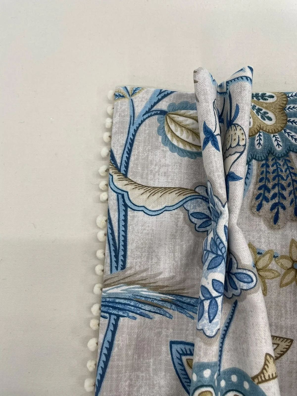 Close-up of a fabric curtain with a blue, beige, and white floral and geometric pattern, decorated with small white pom-poms along the edge.
