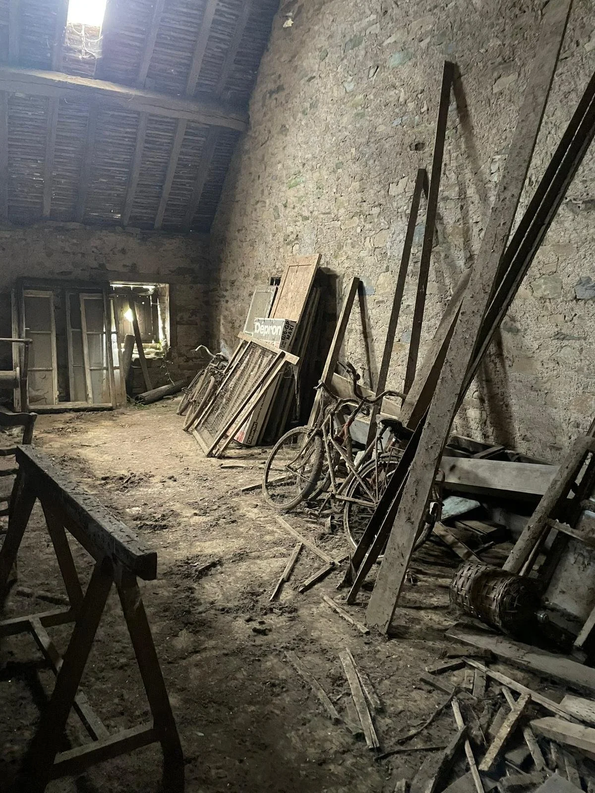 An old barn or storage shed with a dirt floor, cluttered with abandoned bicycles, wooden boards, window frames, and debris, with sunlight streaming through a small opening in the roof.
