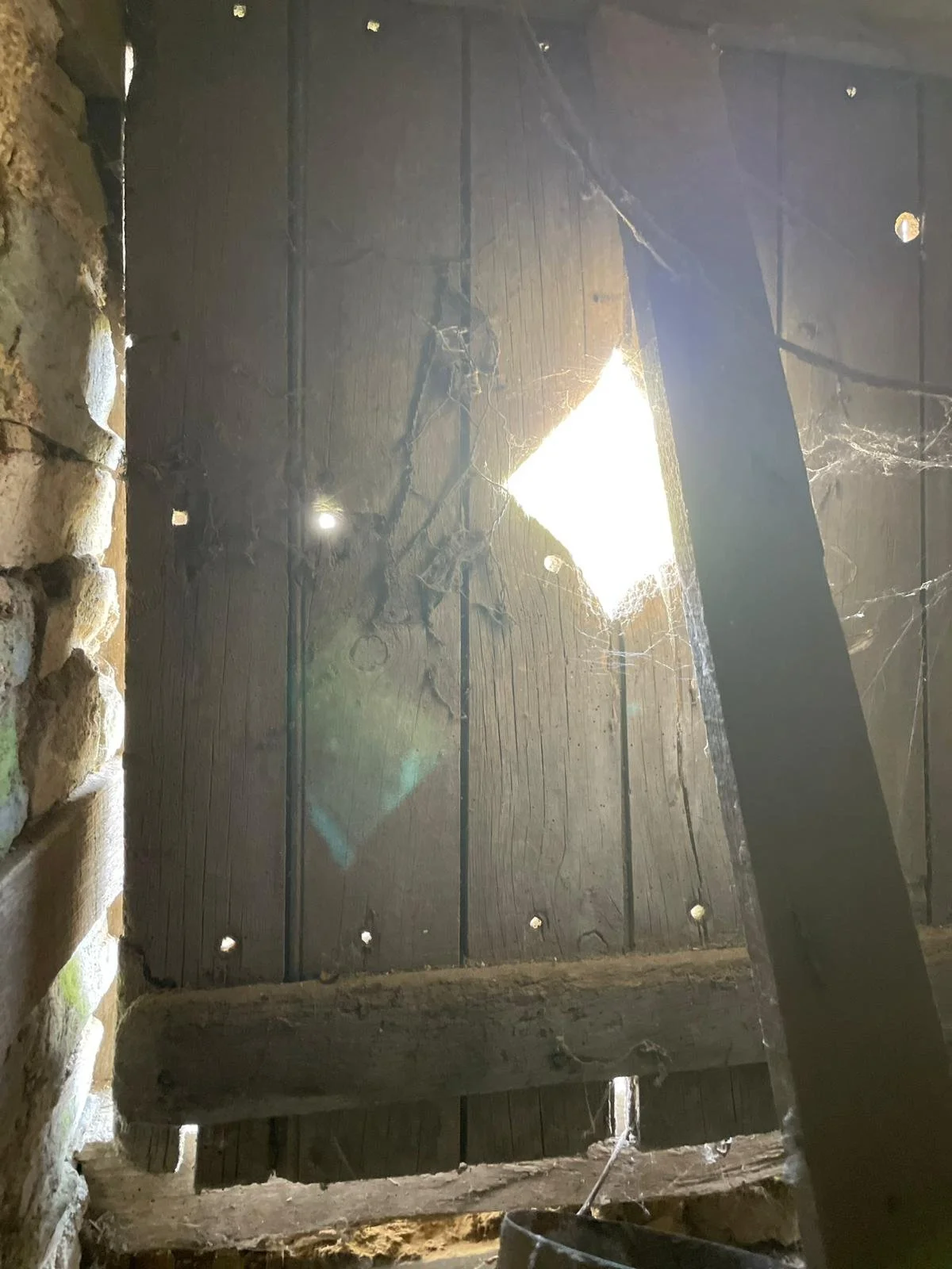 Looking up at a wooden ceiling with sunlight streaming through a small opening, cobwebs hanging, and part of a stone wall on the left.