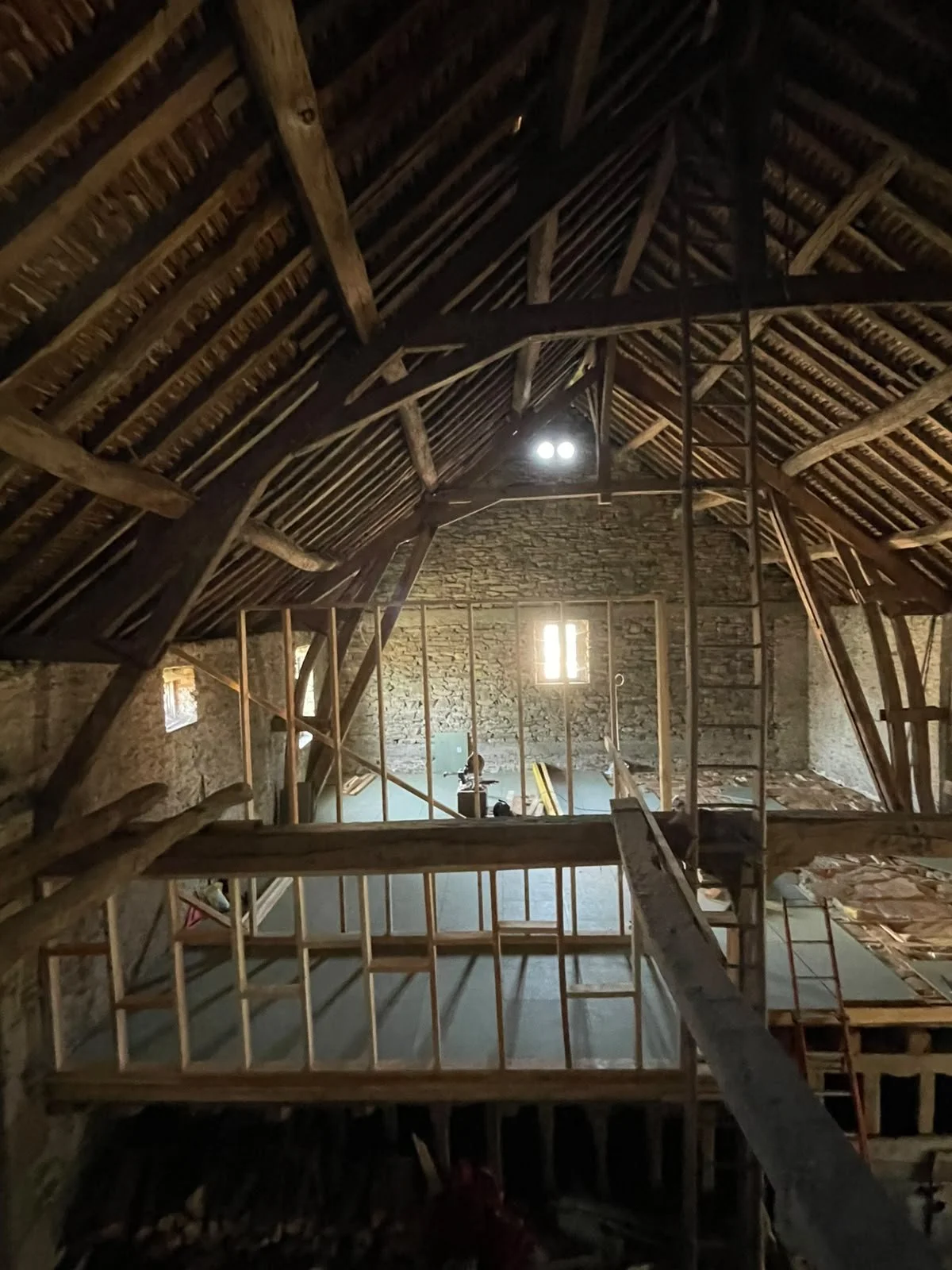Interior of an attic under construction with wooden framing, ladder, and a small window letting in light.