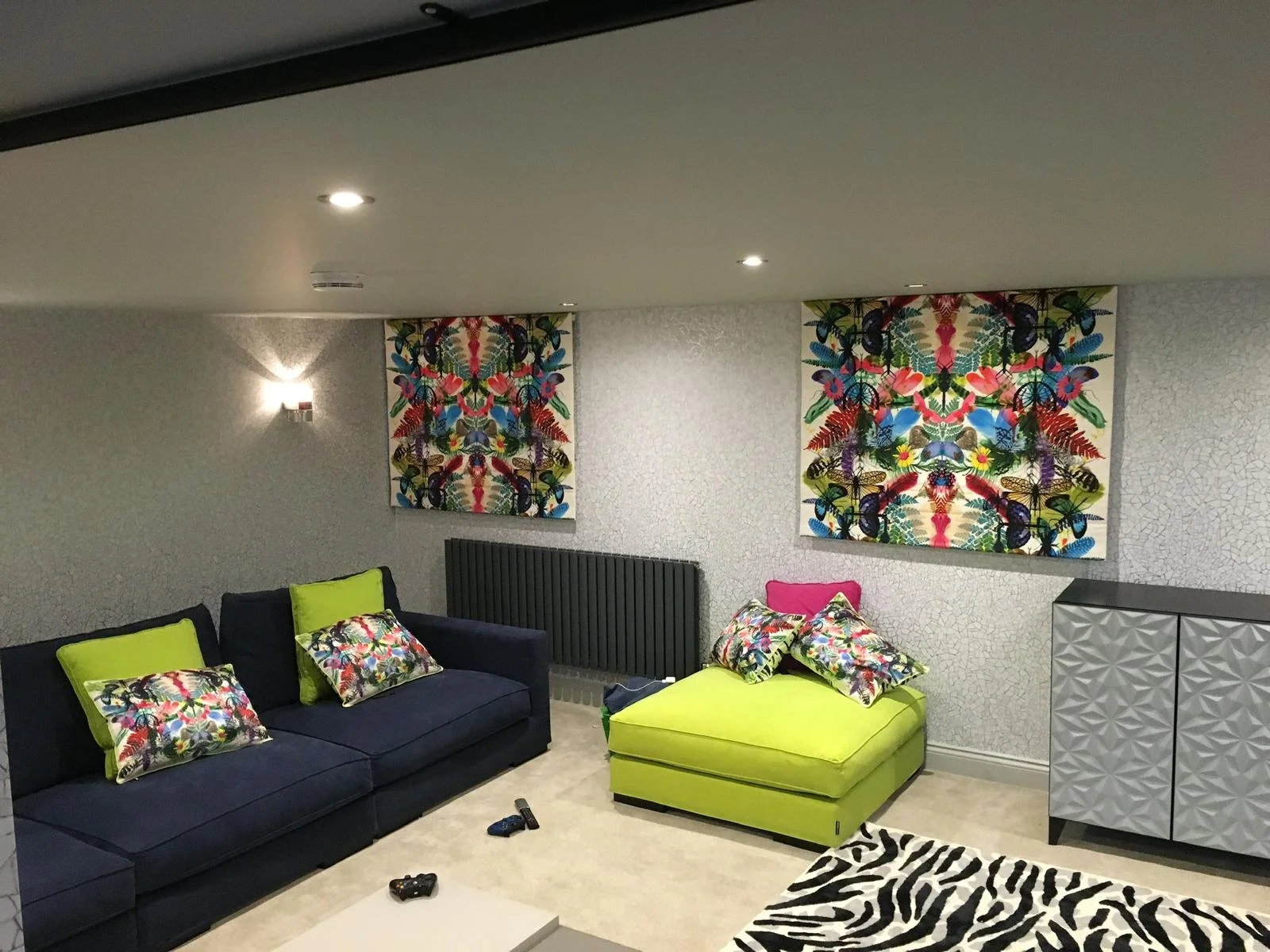 A modern living room with a navy blue sofa and a lime green chaise lounge, both with colorful floral pillows. Two vibrant, symmetrical butterfly and floral patterned artworks hang on a textured white wall, with a black radiator below one. A patterned cabinet and a zebra-striped rug are also visible. Video game controllers rest on the floor.