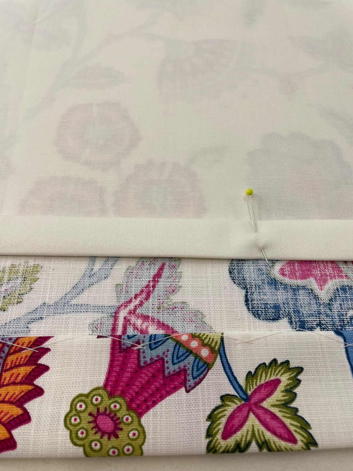 A piece of floral patterned fabric with pink, green, and blue designs underneath a white sewing pattern with a yellow-headed pin securing the fabric and pattern together.