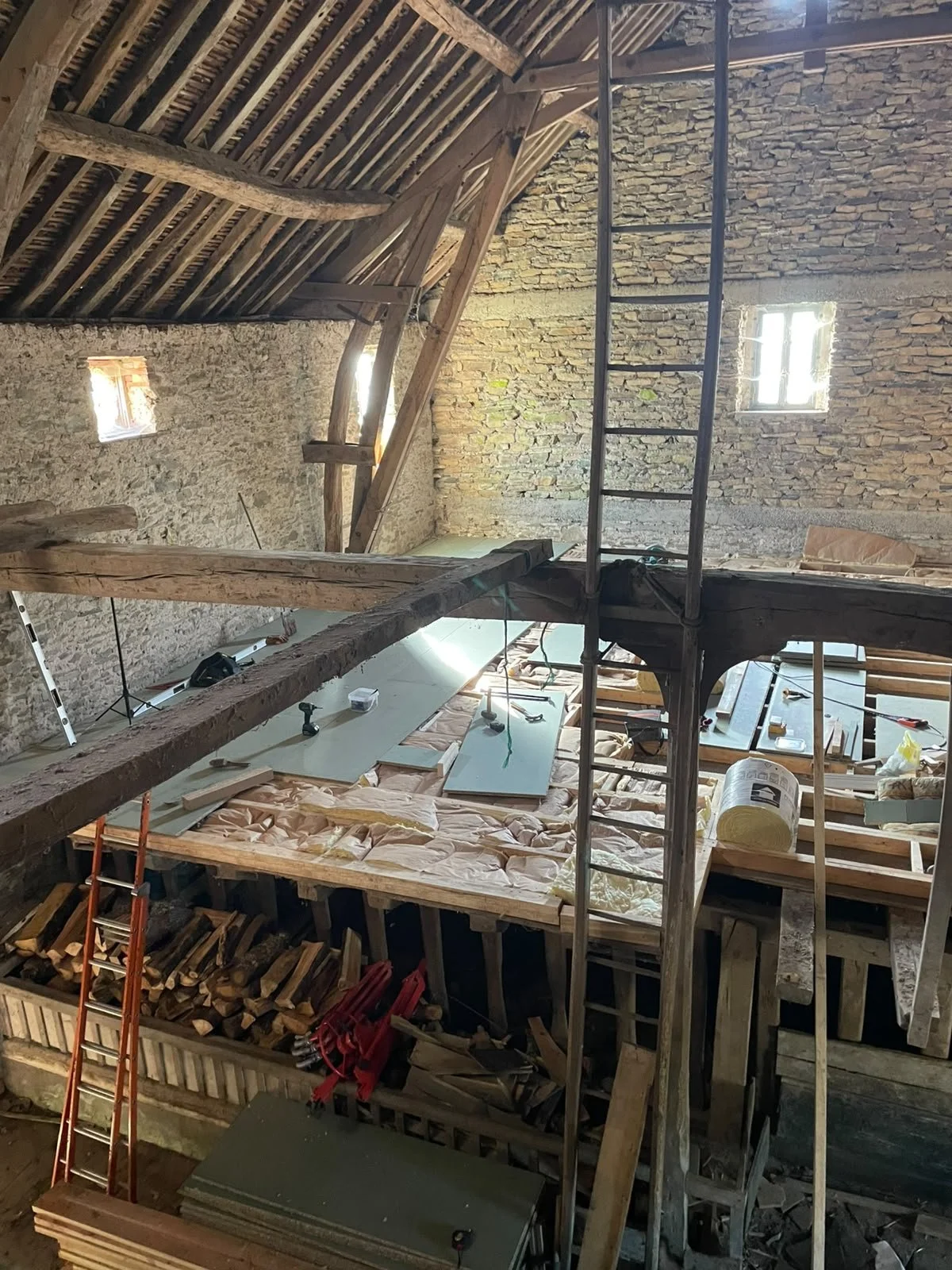 Construction or renovation site inside a building with exposed brick walls, wooden beams, a ladder, construction tools and materials, and insulation on a work surface.