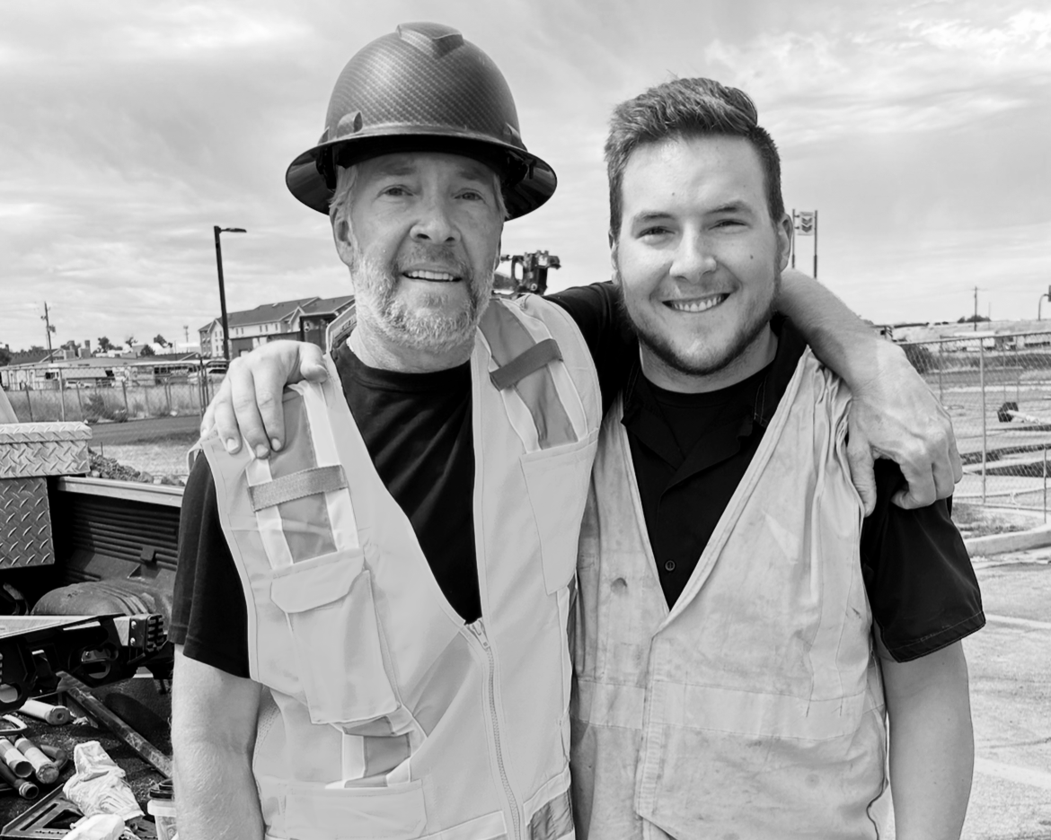 Two men smiling at a construction site, one wearing a hard hat and safety vest, and the other wearing a safety vest with an arm around the other