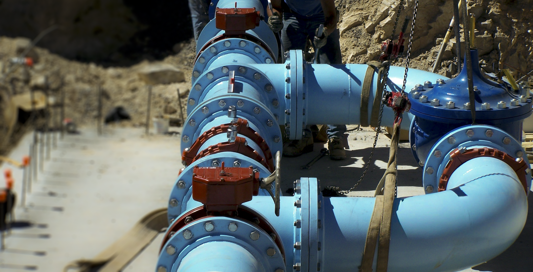 Large blue pipeline with multiple valves being installed at a construction site. Workers in the background are working on the pipeline, surrounded by dirt and construction equipment.