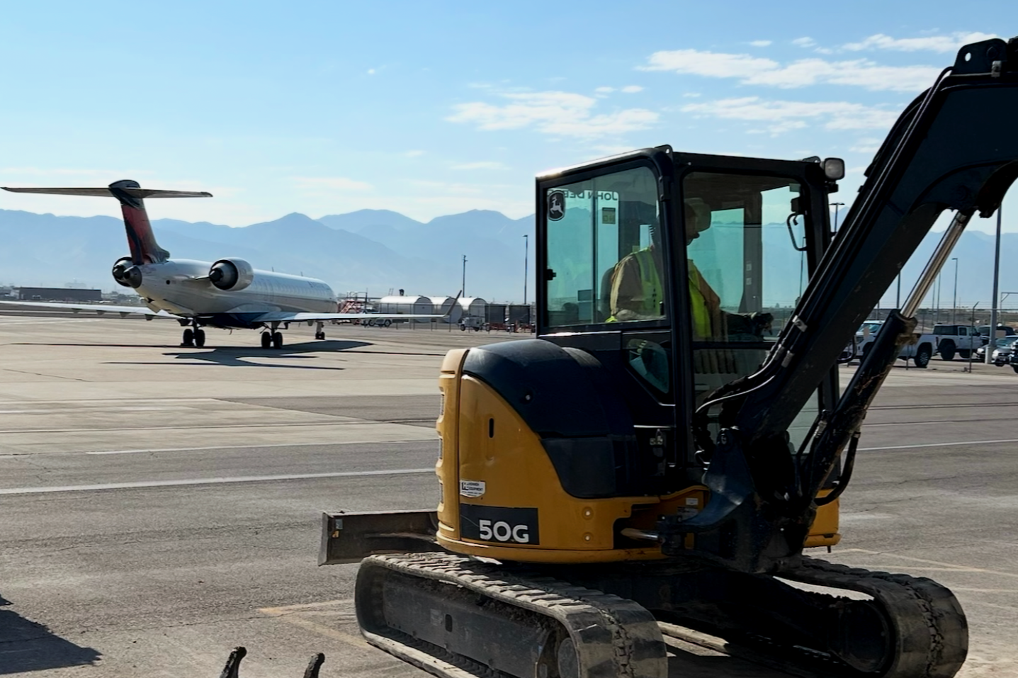 A small yellow excavator with a person inside the cabin, working on a tarmac at an airport. A passenger jet airplane is in the background, along with airport buildings and mountains under a clear blue sky.