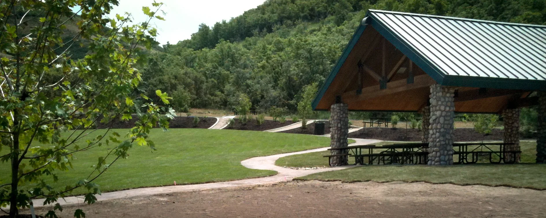 A park with a pavilion supported by stone pillars, surrounded by a grassy lawn and trees, with a winding pathway leading into a wooded area.