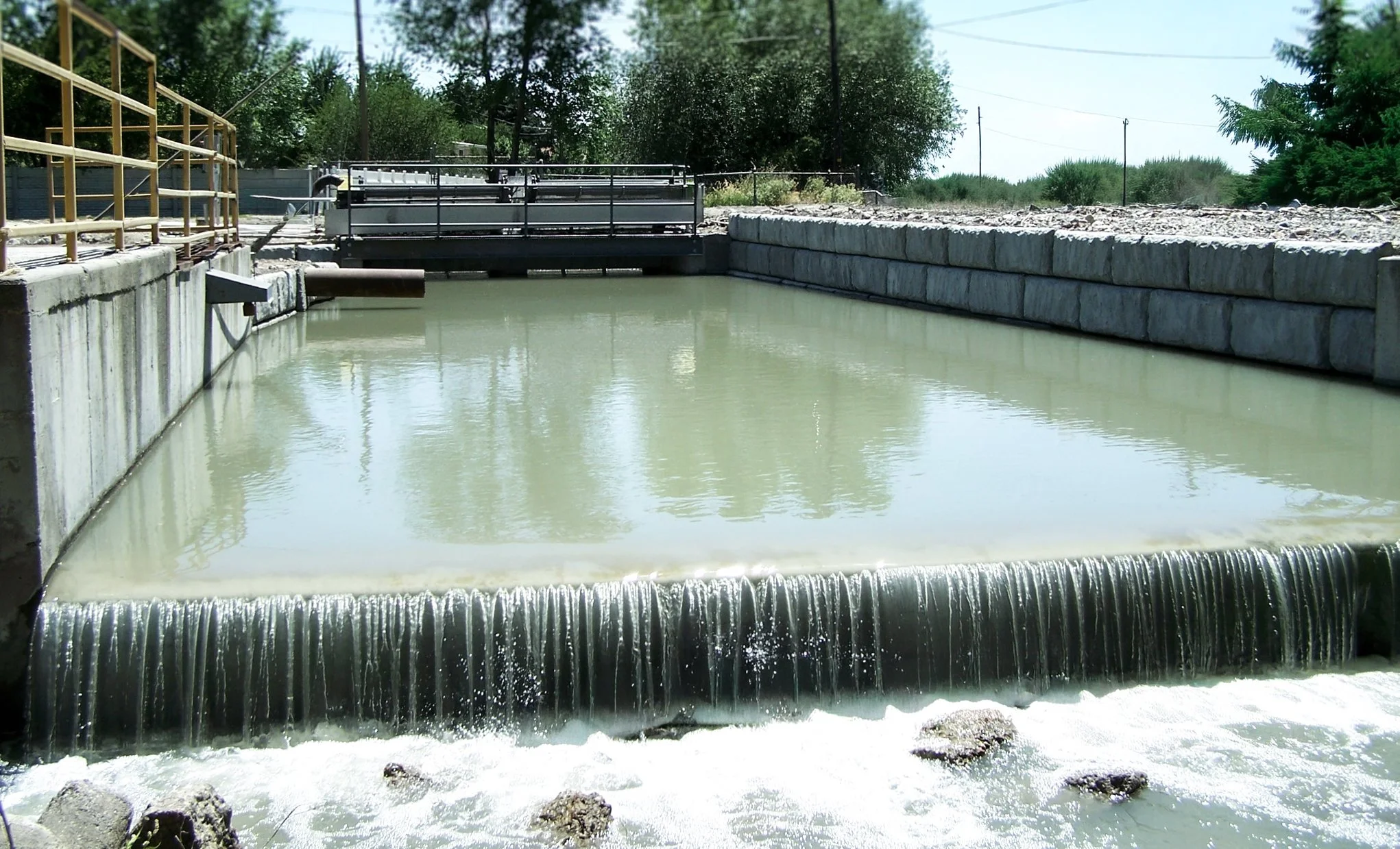 A canal or waterway with a weir or small dam, surrounded by concrete walls and metal railings, with water flowing over the edge.