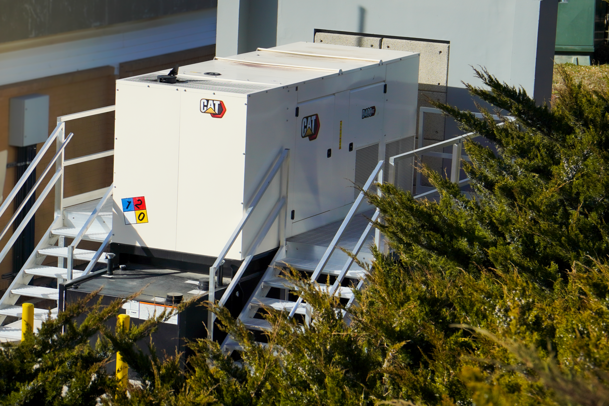 Large white industrial generator with CAT logo, positioned on an elevated platform with metal stairs, surrounded by green shrubbery and a building wall in the background.