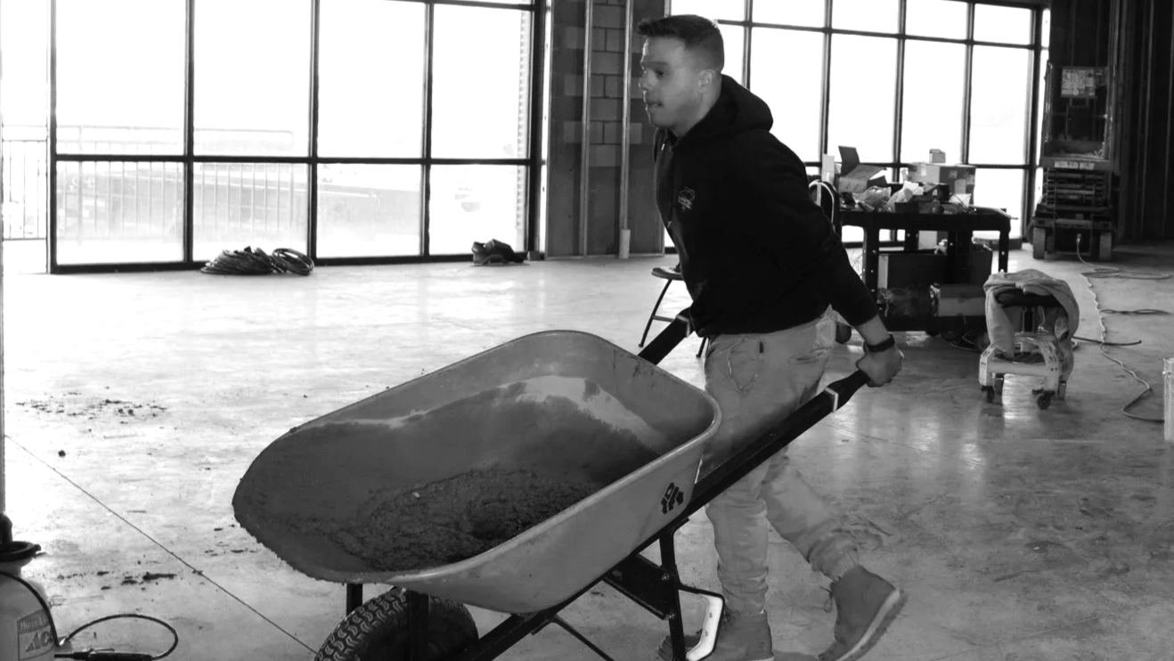 A man in a hoodie and jeans pushing a wheelbarrow with concrete inside, working in an unfinished indoor space with large windows and construction tools around.