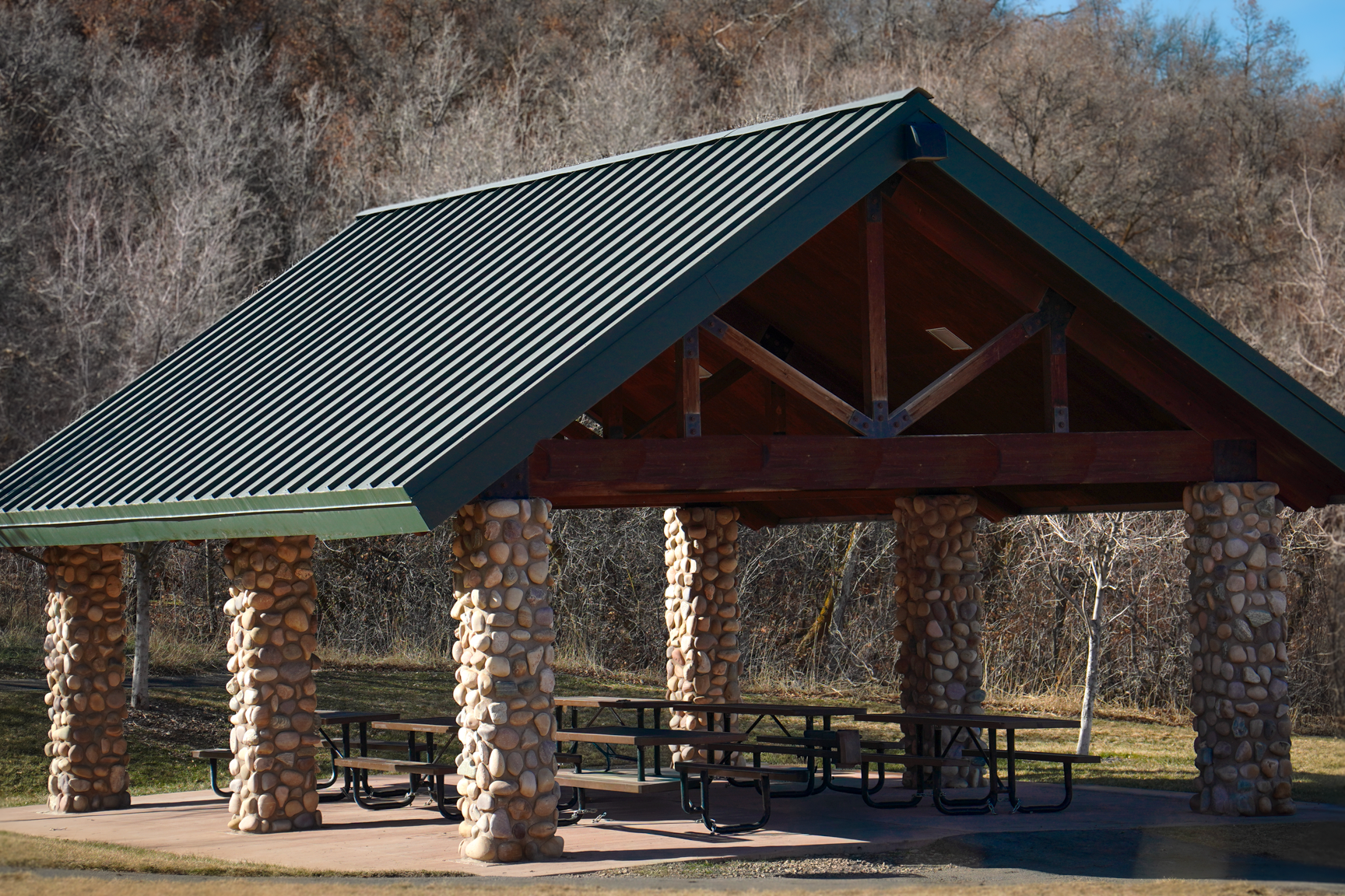 An outdoor picnic shelter with stone columns, a metal gabled roof, and picnic tables underneath, set against a wooded area with leafless trees.