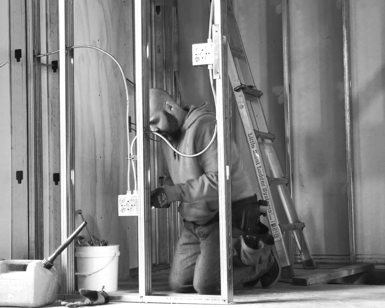 A man with a beard kneels inside a framed metal wall during electrical wiring work, with tools, a ladder, and construction materials around him.